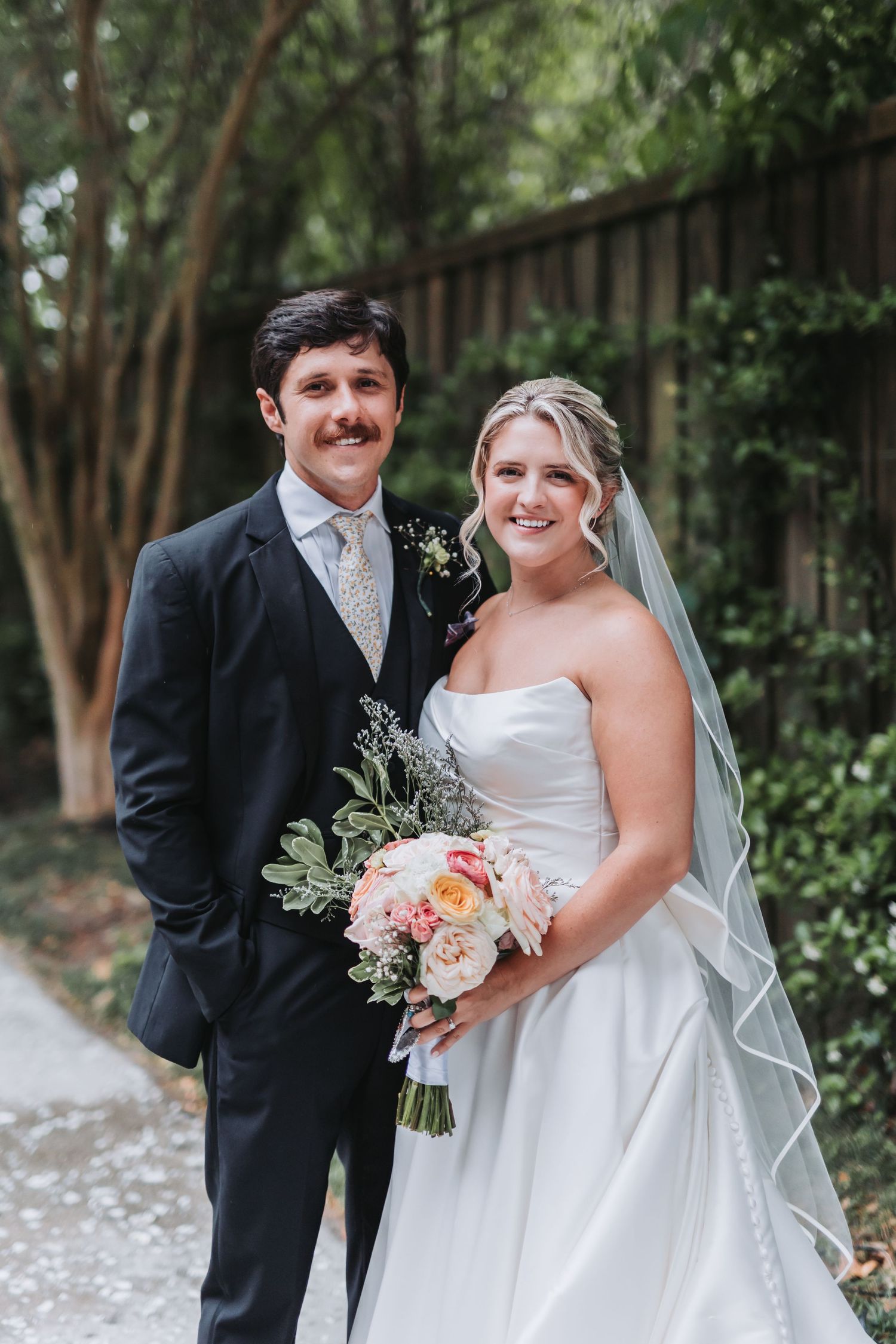 Smiling bride and groom pose together outdoors, bride holding a pastel bouquet of roses and greenery.