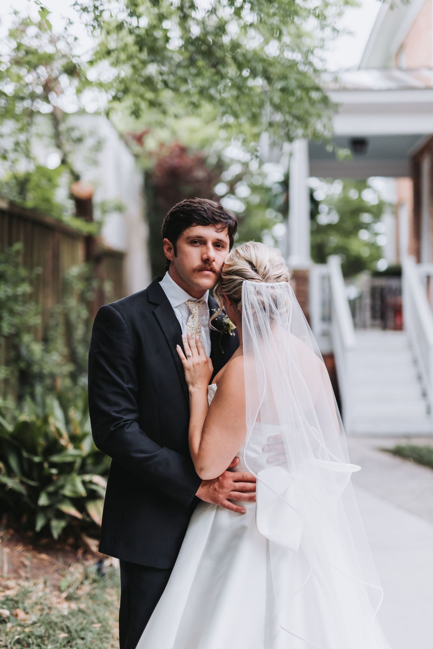Groom in black suit embraces bride in white gown with veil in lush garden setting.