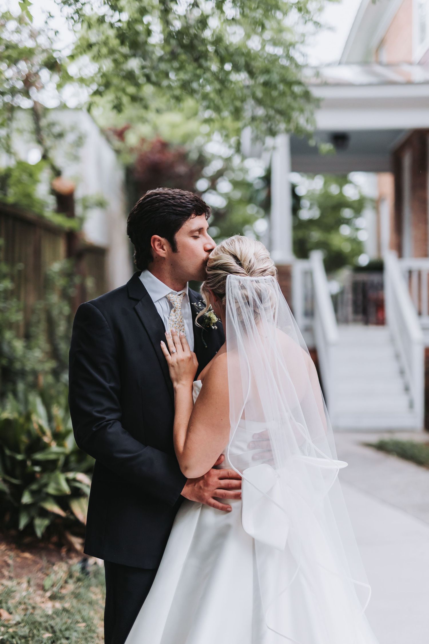 Bride in white gown and veil shares romantic kiss with groom in black suit in lush garden setting.