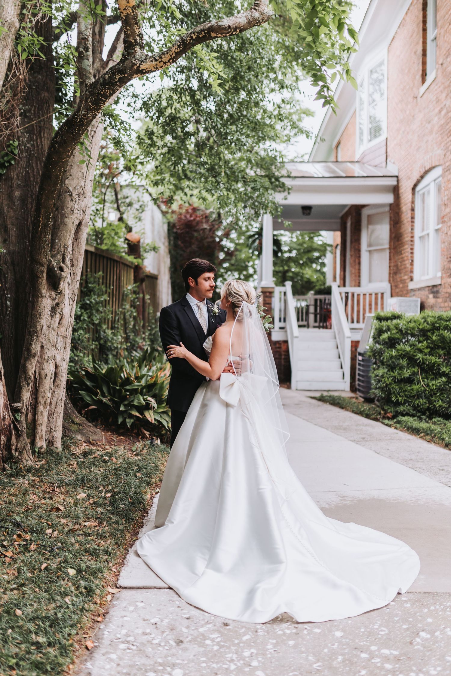 Bride in long white gown with veil and groom embrace on garden path beside brick building.