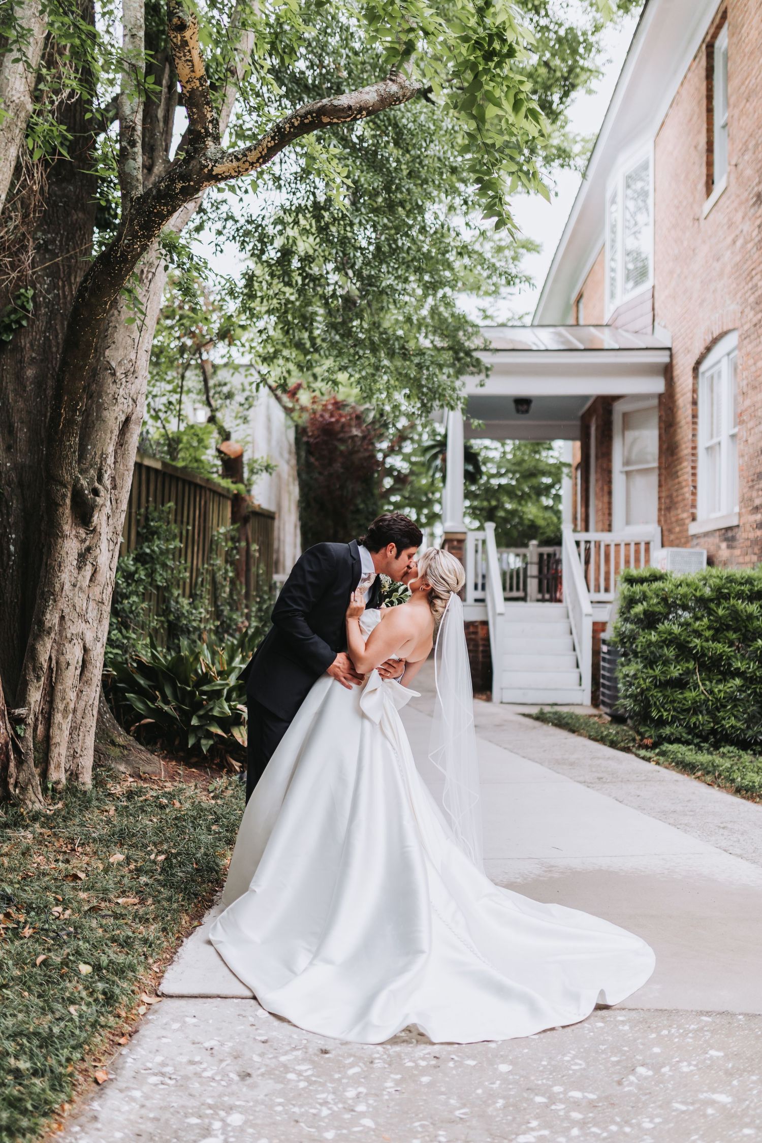 Groom dips bride in white gown with long train for romantic kiss on tree-lined walkway.