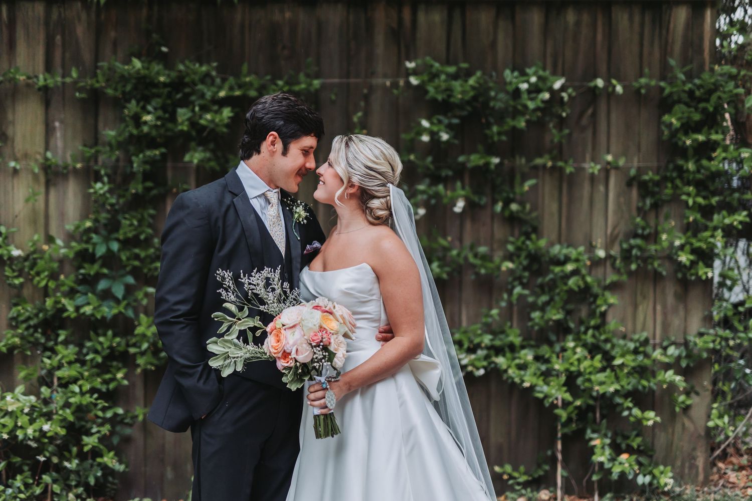 Bride and groom share romantic moment in front of ivy-covered fence, bride holds bouquet.
