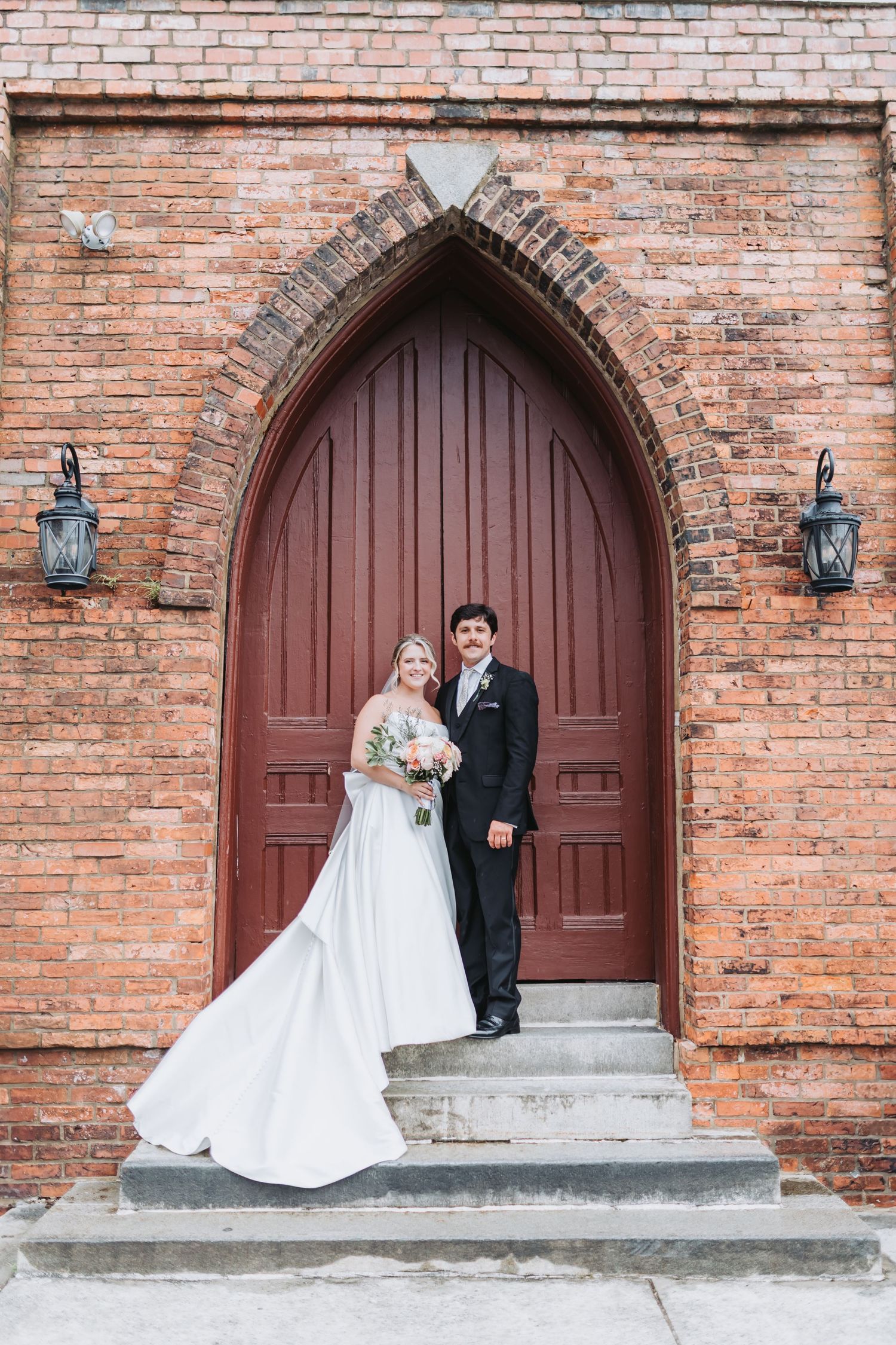Bride and groom posing on church steps before a grand arched red wooden door with brick wall backdrop.