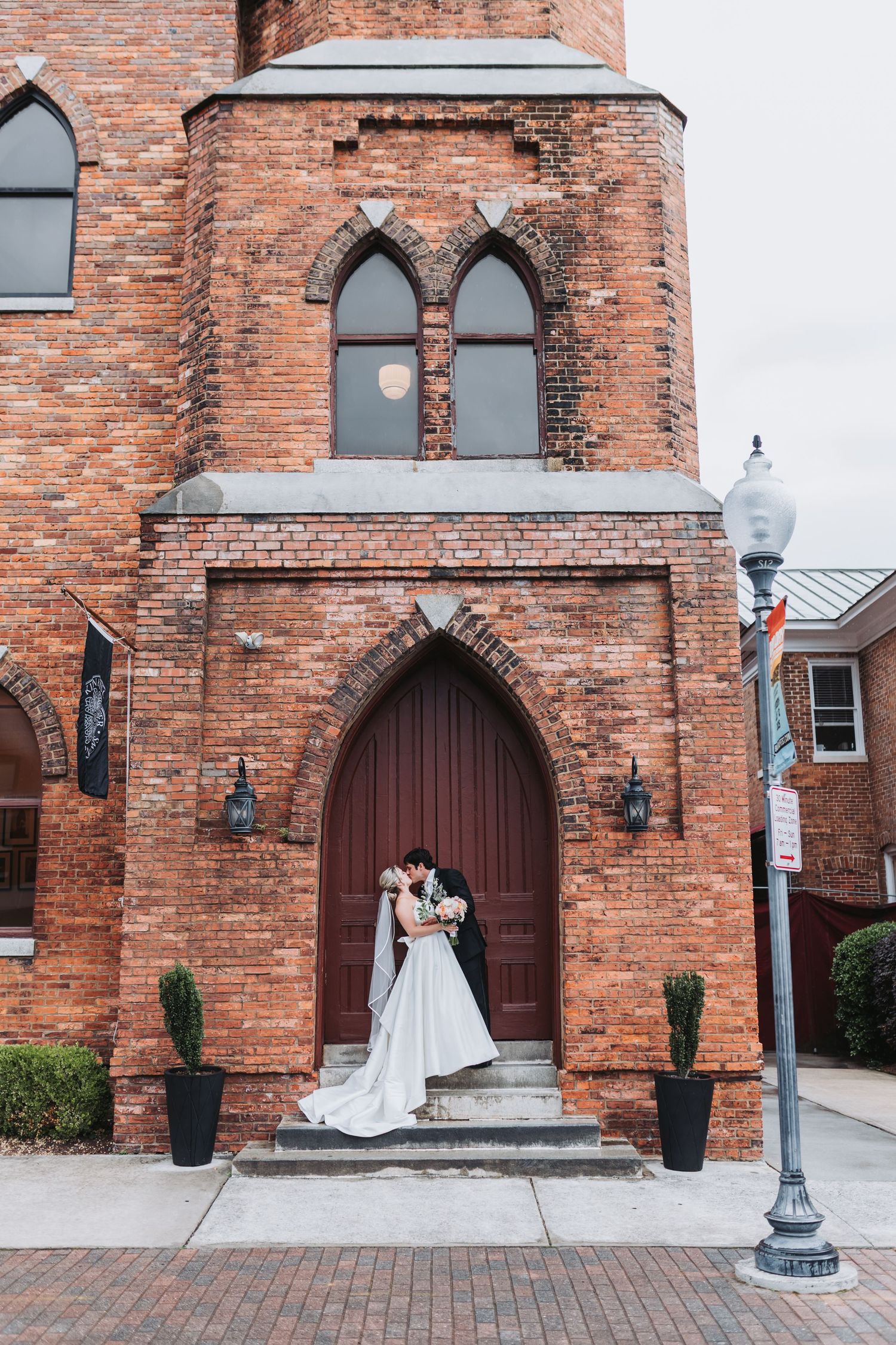 Bride and groom kiss in front of ornate red brick church with arched wooden doors.