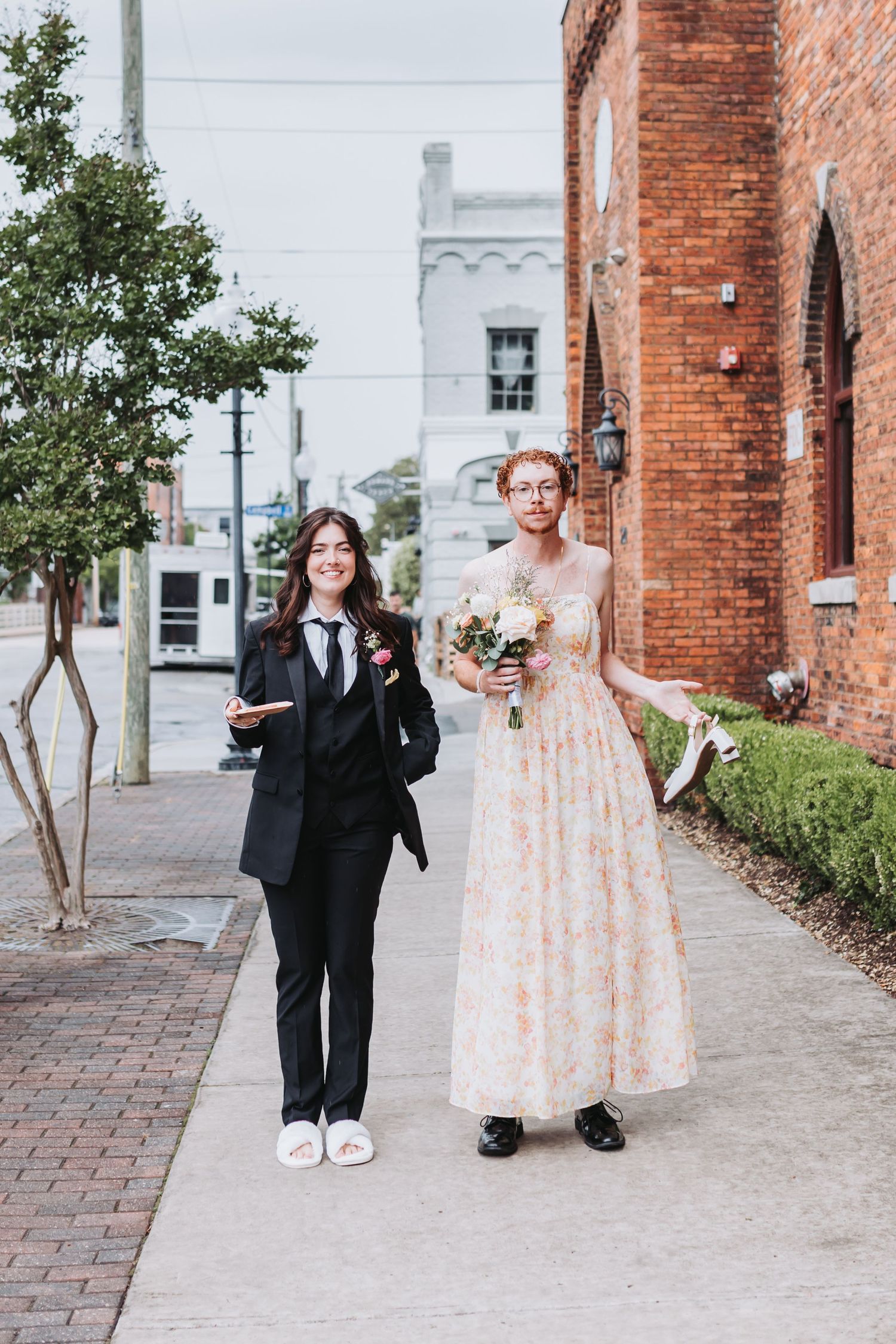 Two brides walking outside brick building, one in black suit with pink boutonniere, other in floral gown holding bouquet.