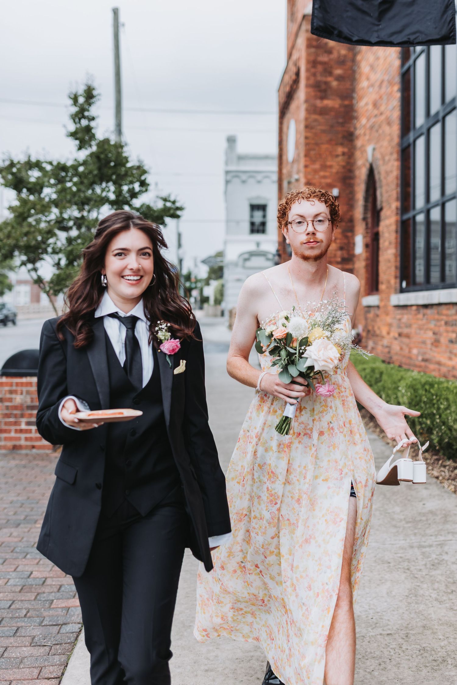 Bridesmaid in black tuxedo walks with groomsman in floral dress holding bouquet outside.