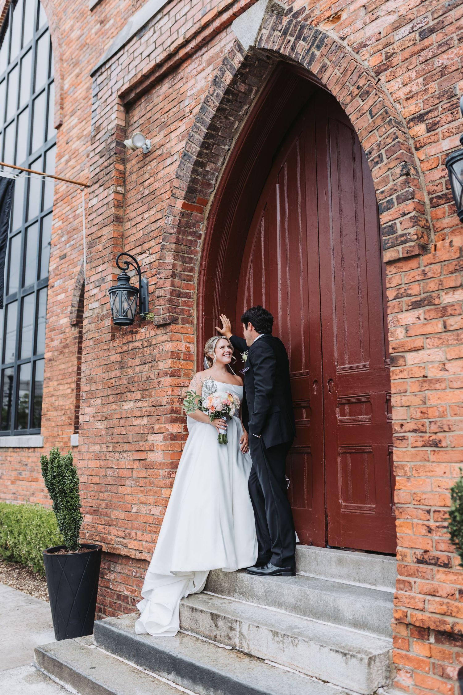 Bride and groom posing at a red arched door of a rustic brick wedding venue.