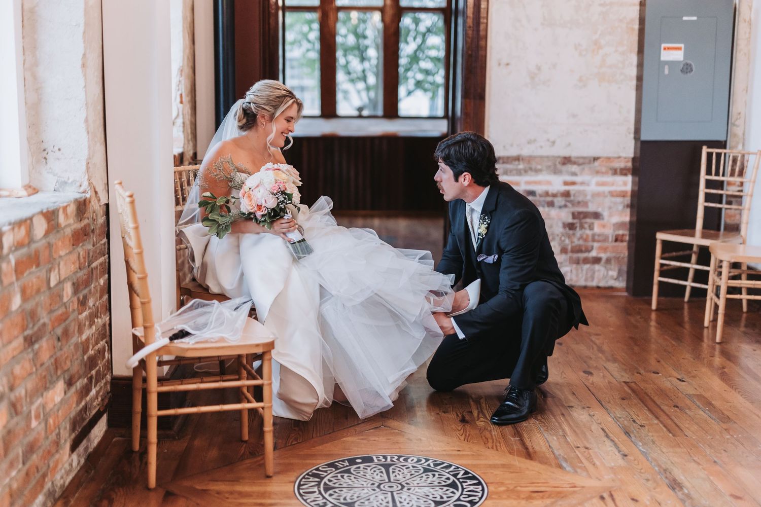 Groom kneels before seated bride in rustic wood-floored venue adjusting her wedding gown train.