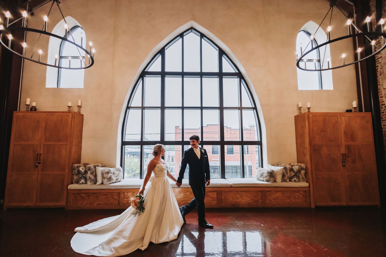 Bride and groom hold hands in elegant chapel with arched gothic window and chandelier lighting.