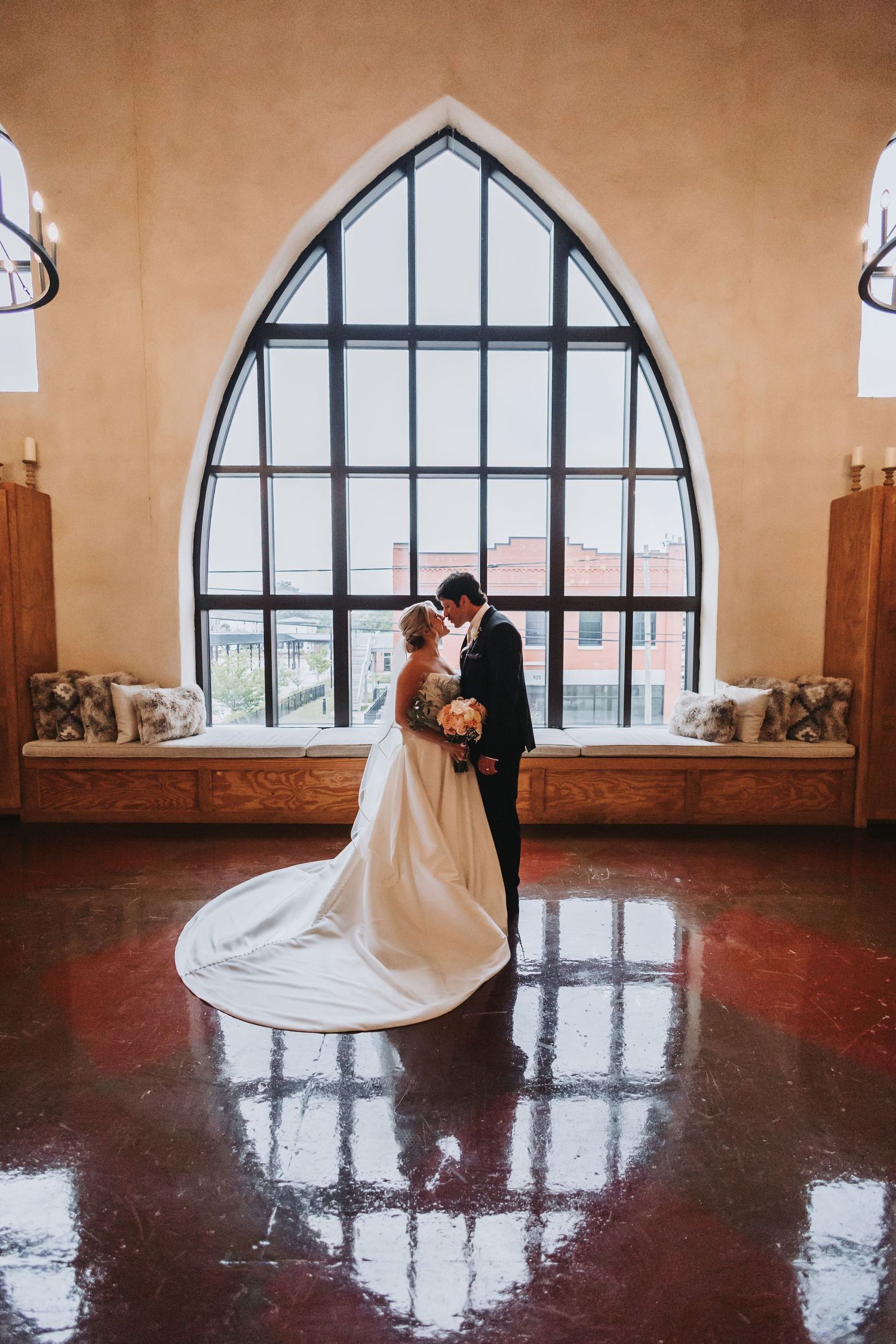 Bride and groom share intimate moment before dramatic arched window in elegant wedding venue interior.