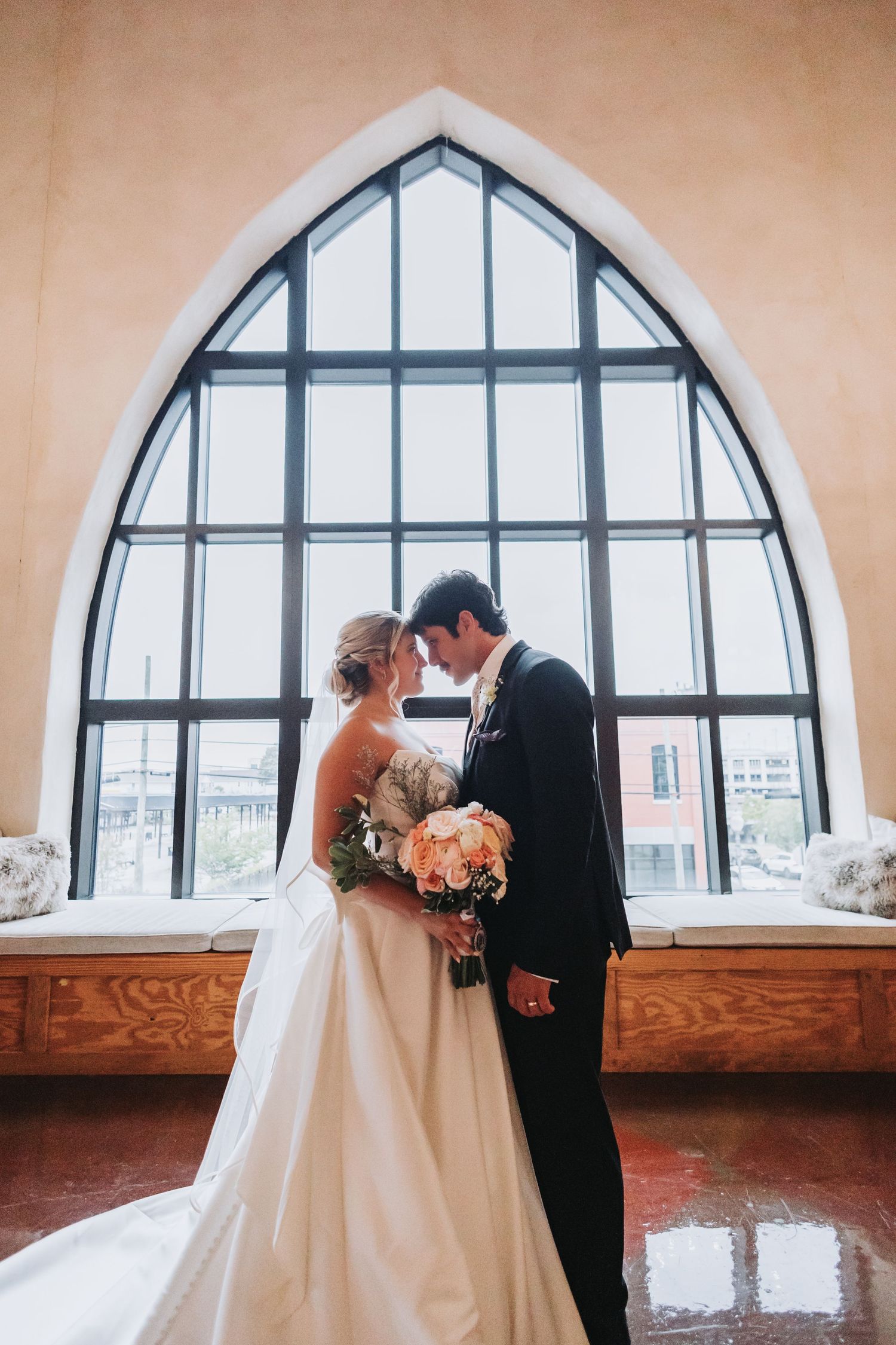 Bride and groom sharing romantic moment before large Gothic arched window holding pink wedding bouquet.