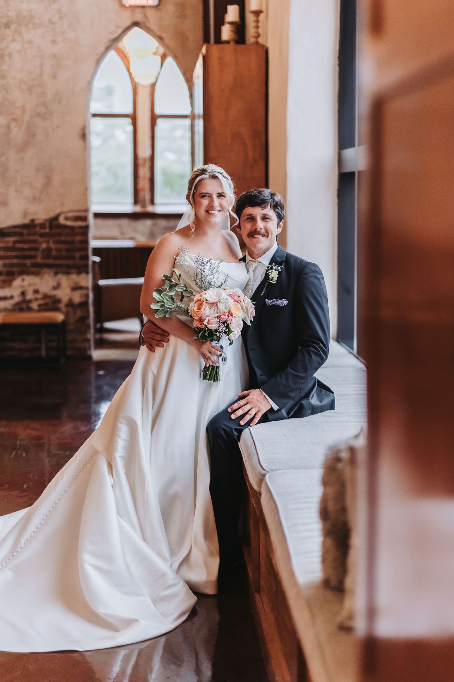 Bride and groom with mustache pose together inside rustic brick venue by arched window with bouquet.