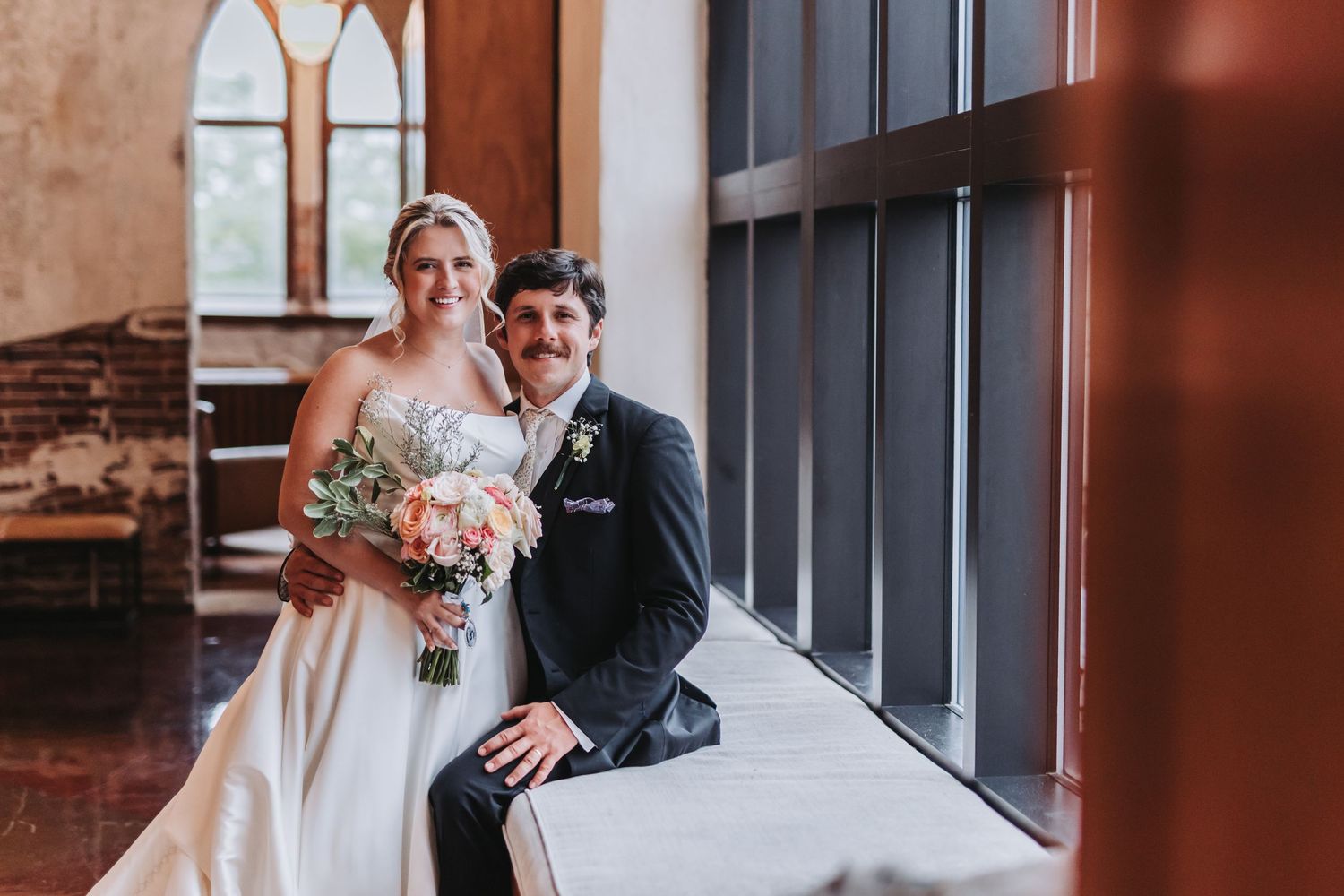 Bride and groom pose together near window inside brick church after wedding ceremony.