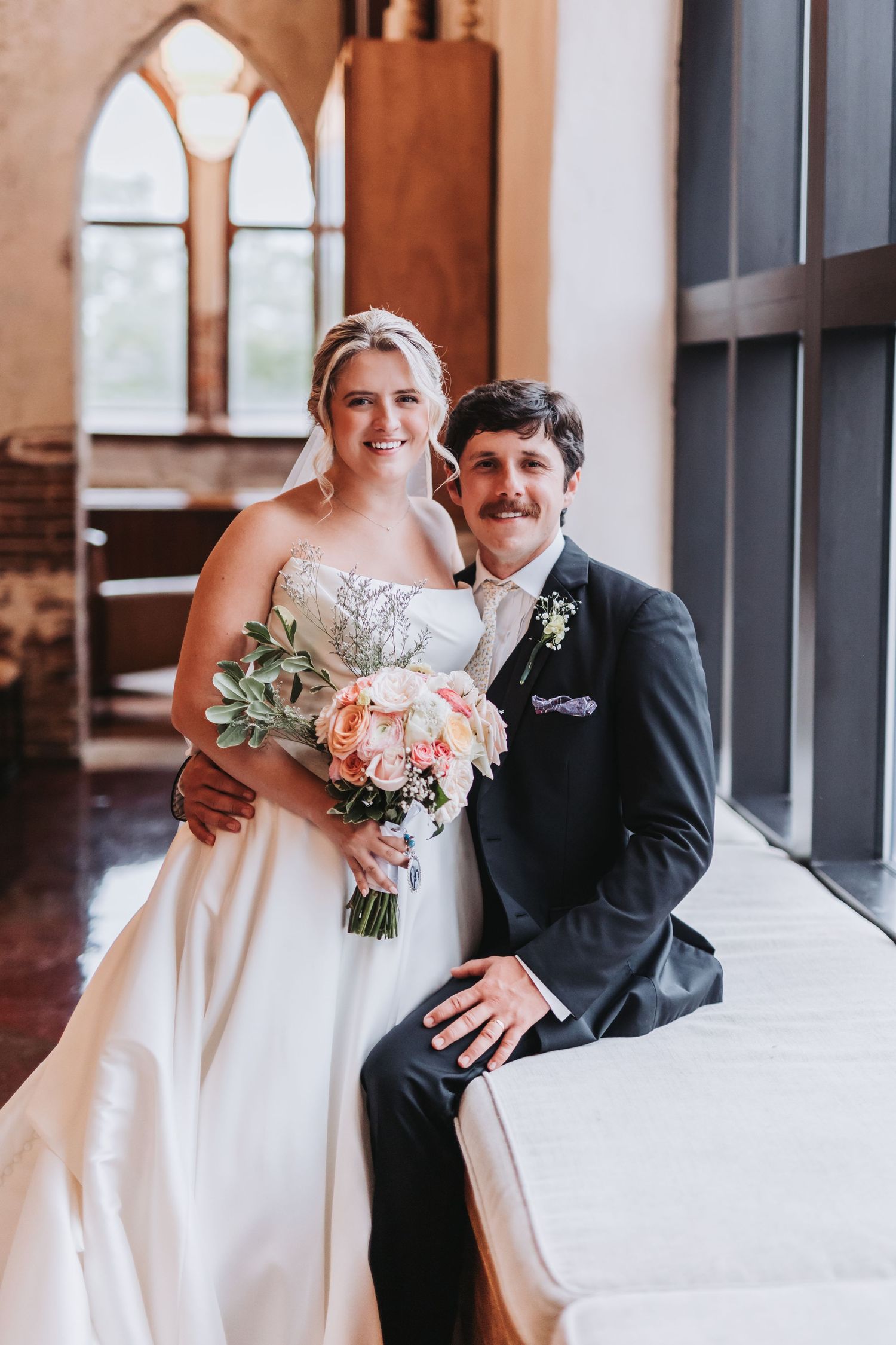 Smiling bride and groom posing together inside elegant church venue with arched windows and natural light.