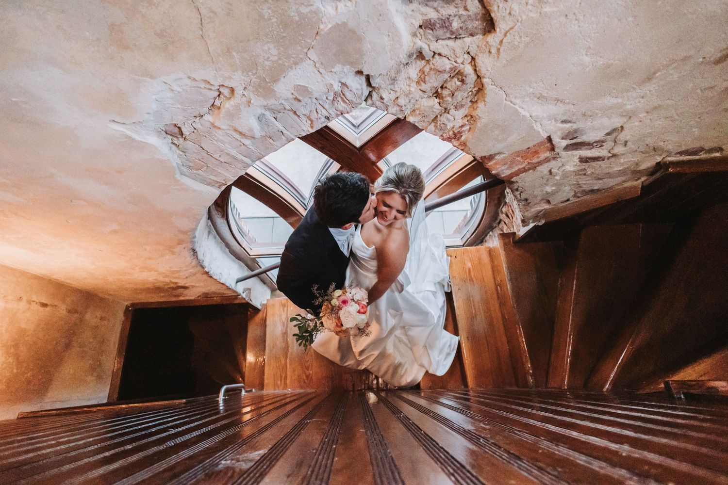 Bride and groom share romantic moment in unique spiral wooden staircase viewed from above.
