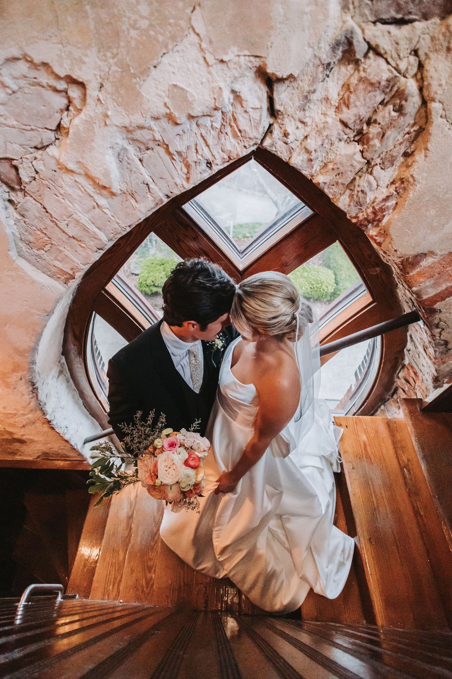 Bride and groom share a kiss on spiral wooden staircase beneath a round window.