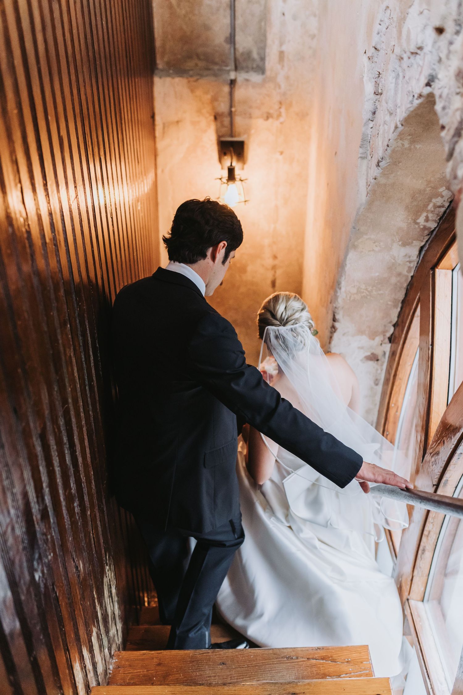 Groom guides bride in white gown and veil up narrow warmly lit interior staircase.