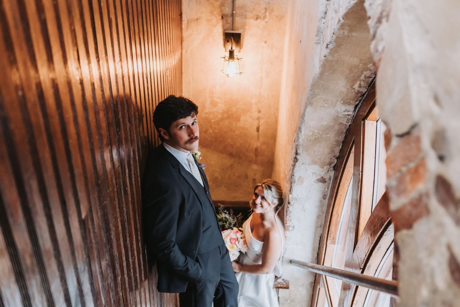 Bride and groom pose in rustic stairwell with exposed brick wall and vintage Edison bulb lighting.