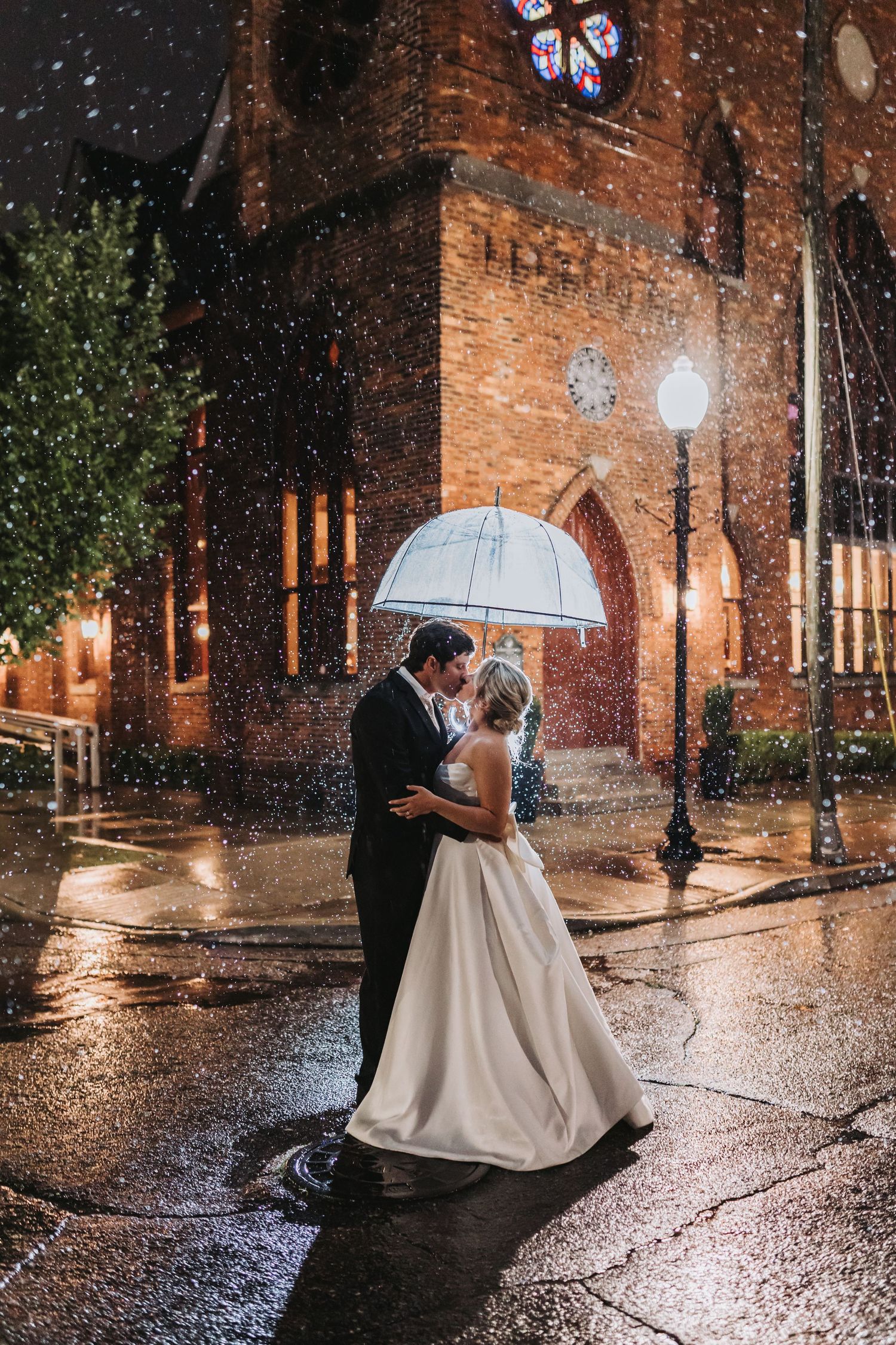 Bride and groom share a romantic kiss under a clear umbrella in the rain outside a lit brick church at night.