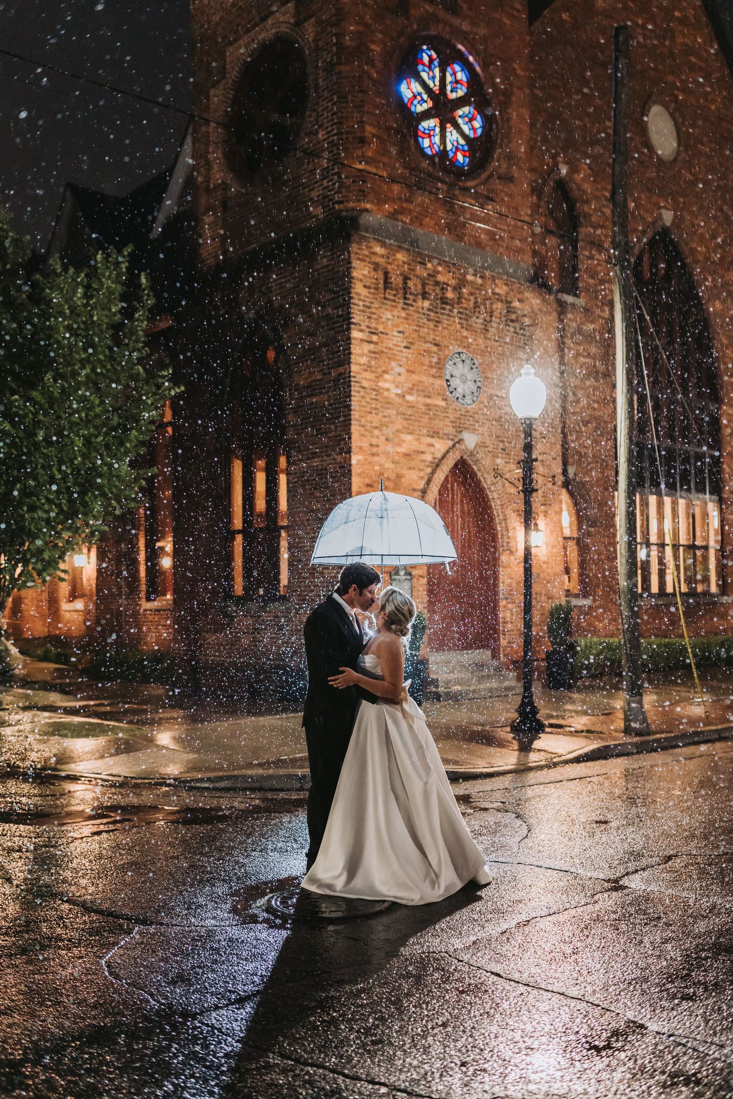 Bride and groom kissing under clear umbrella in rain outside lit brick church at night.