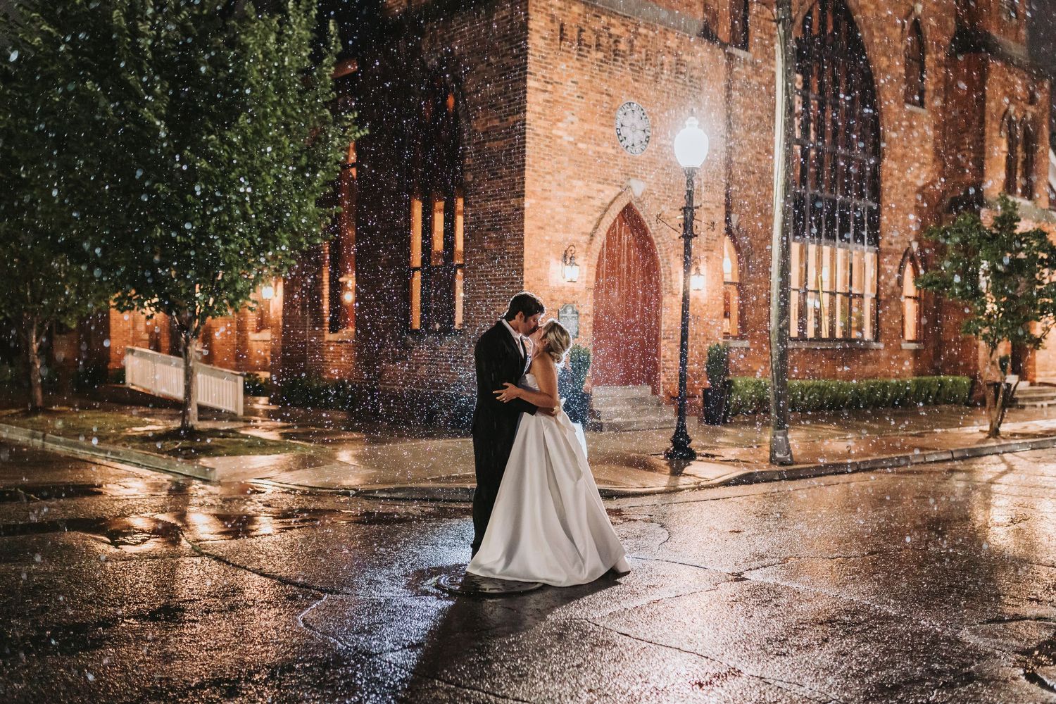 A bride and groom share a romantic embrace in the rain outside a beautifully lit brick church at night.