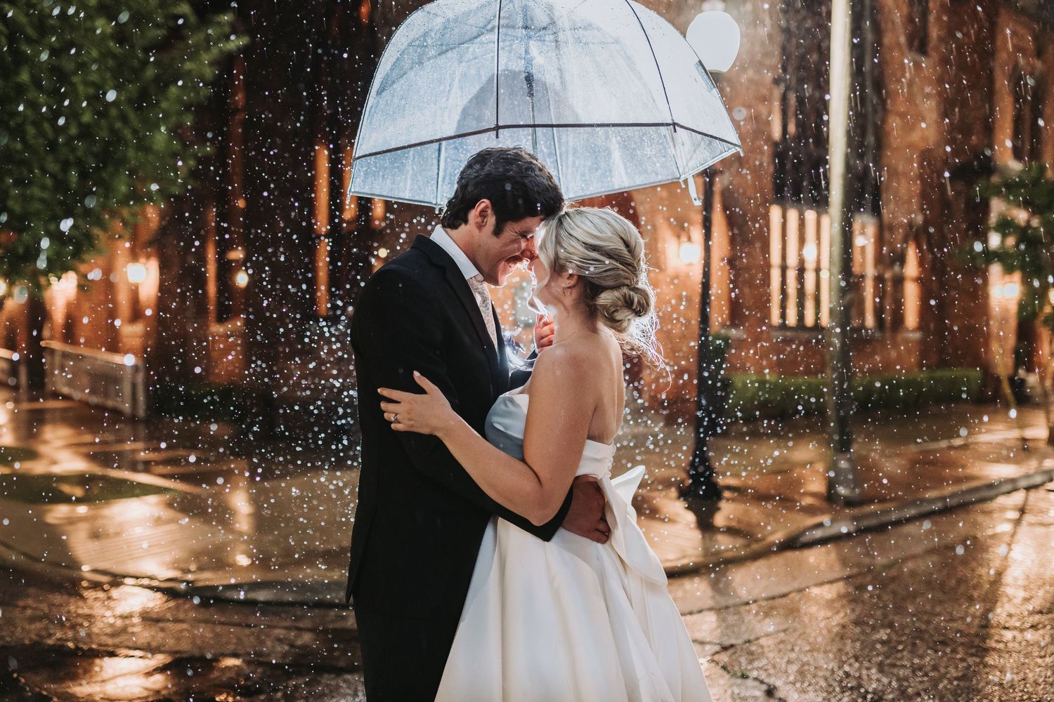 Bride and groom share romantic kiss under clear umbrella in rain on city street at night.