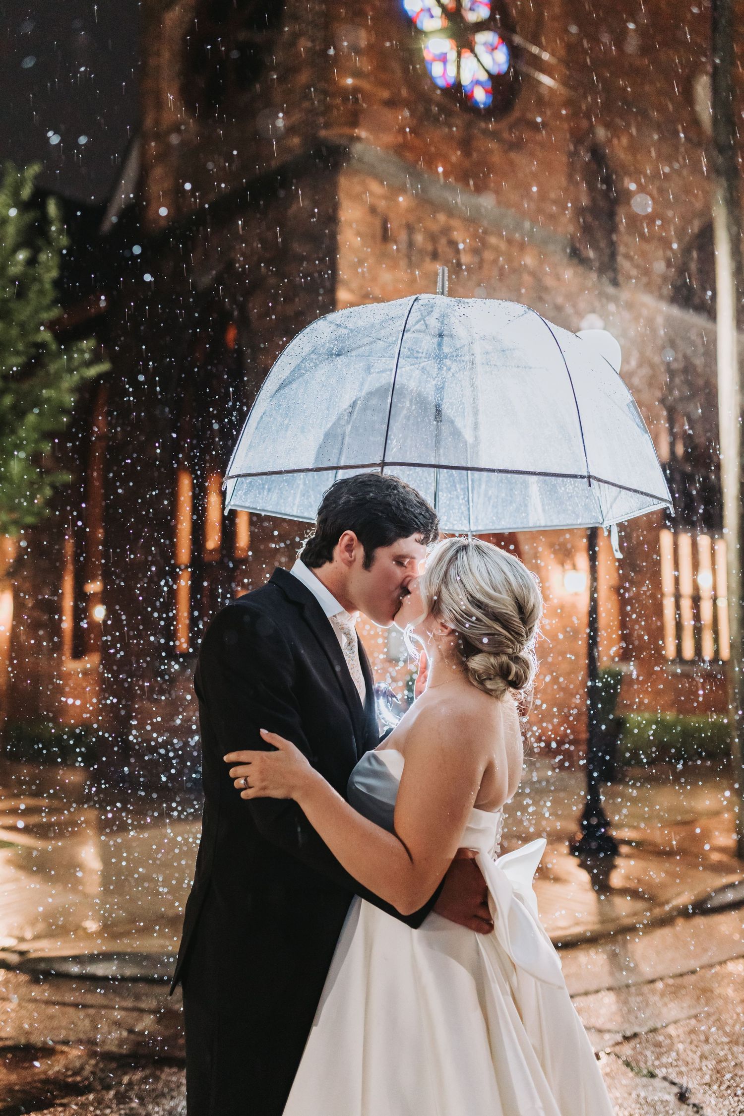 Bride and groom share a romantic kiss under a clear umbrella in the rain outside an illuminated church at night.
