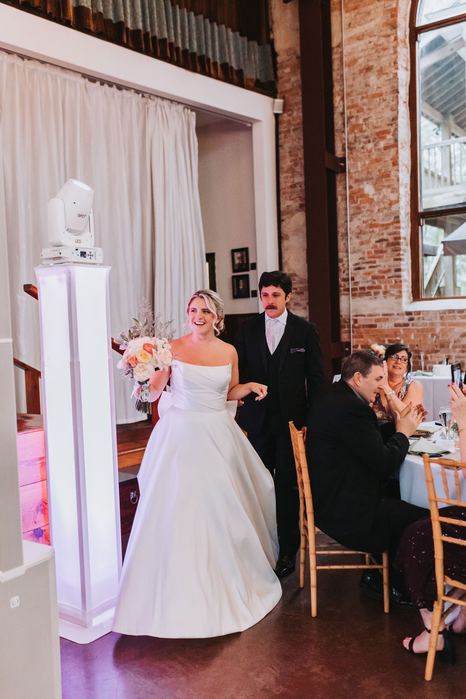 Bride and groom making grand entrance into rustic brick reception hall as guests applaud their arrival.