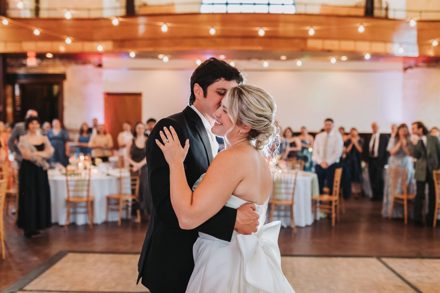 Bride and groom share first dance kiss on ballroom floor surrounded by guests and string lights.