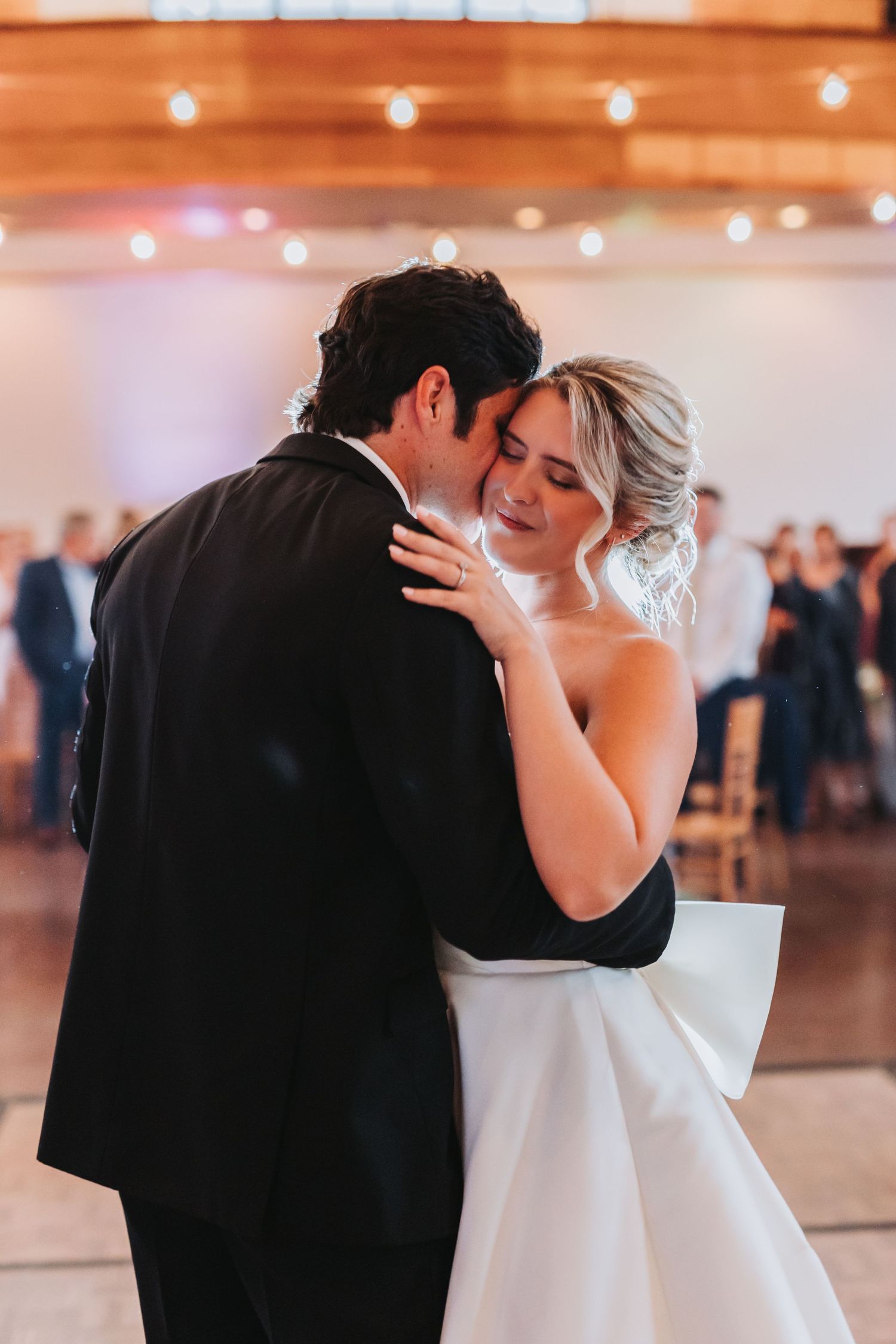 Bride and groom share their first dance, embracing closely under warm string lights at their wedding reception.