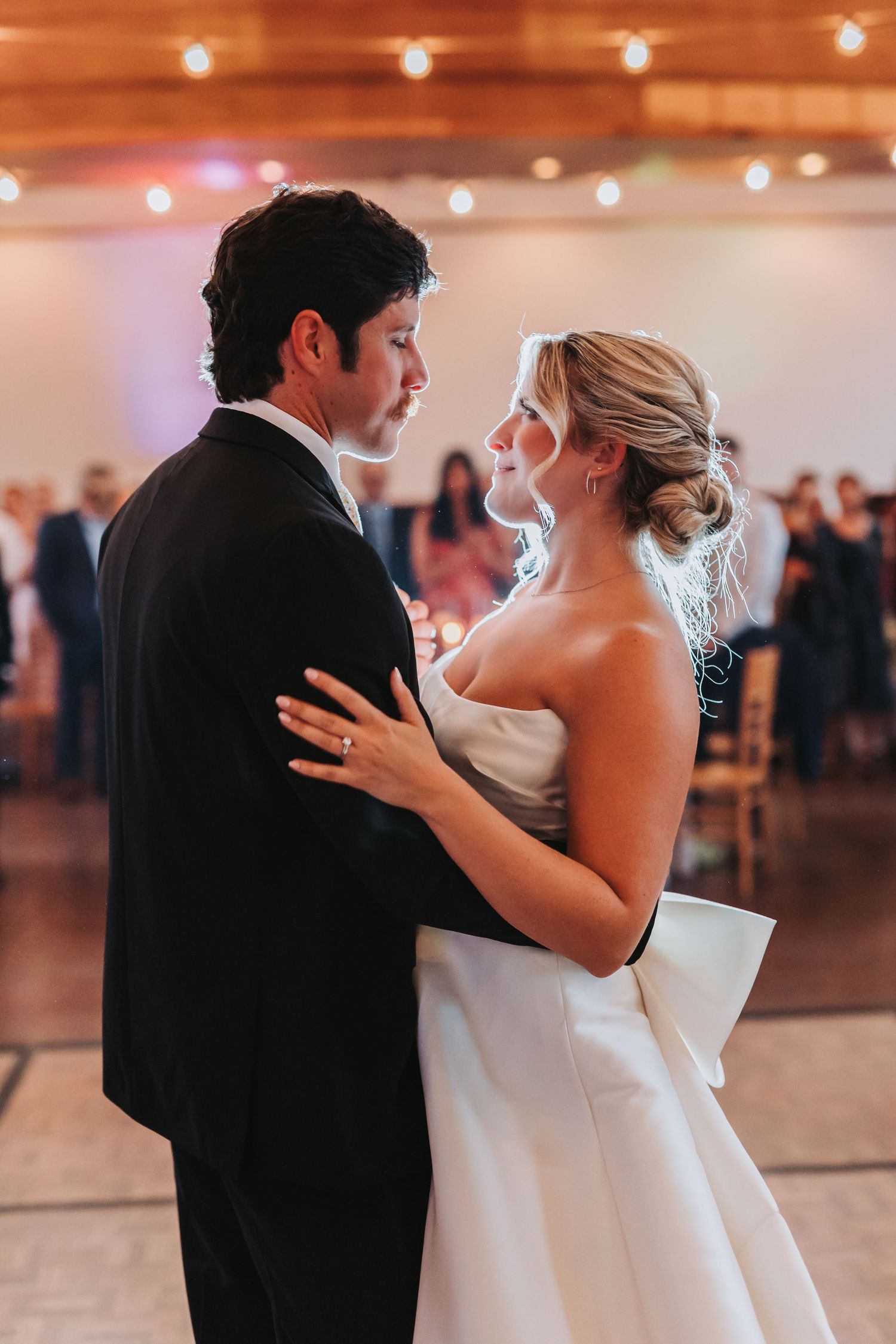 Bride and groom share their first dance, gazing into each other's eyes in a warmly lit wedding reception hall.