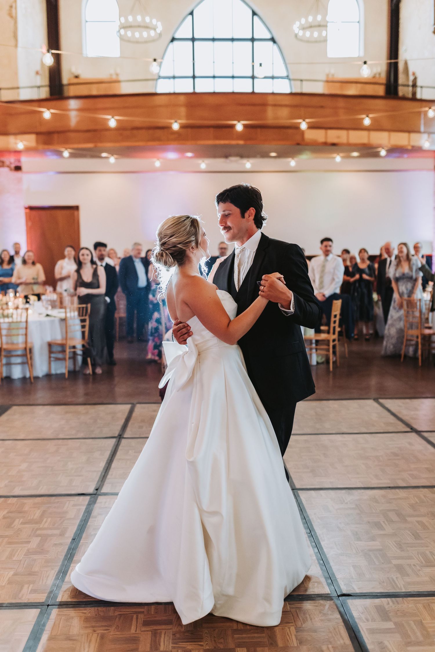 Bride and groom share their first dance in an elegant wedding reception hall with string lights and guests watching.