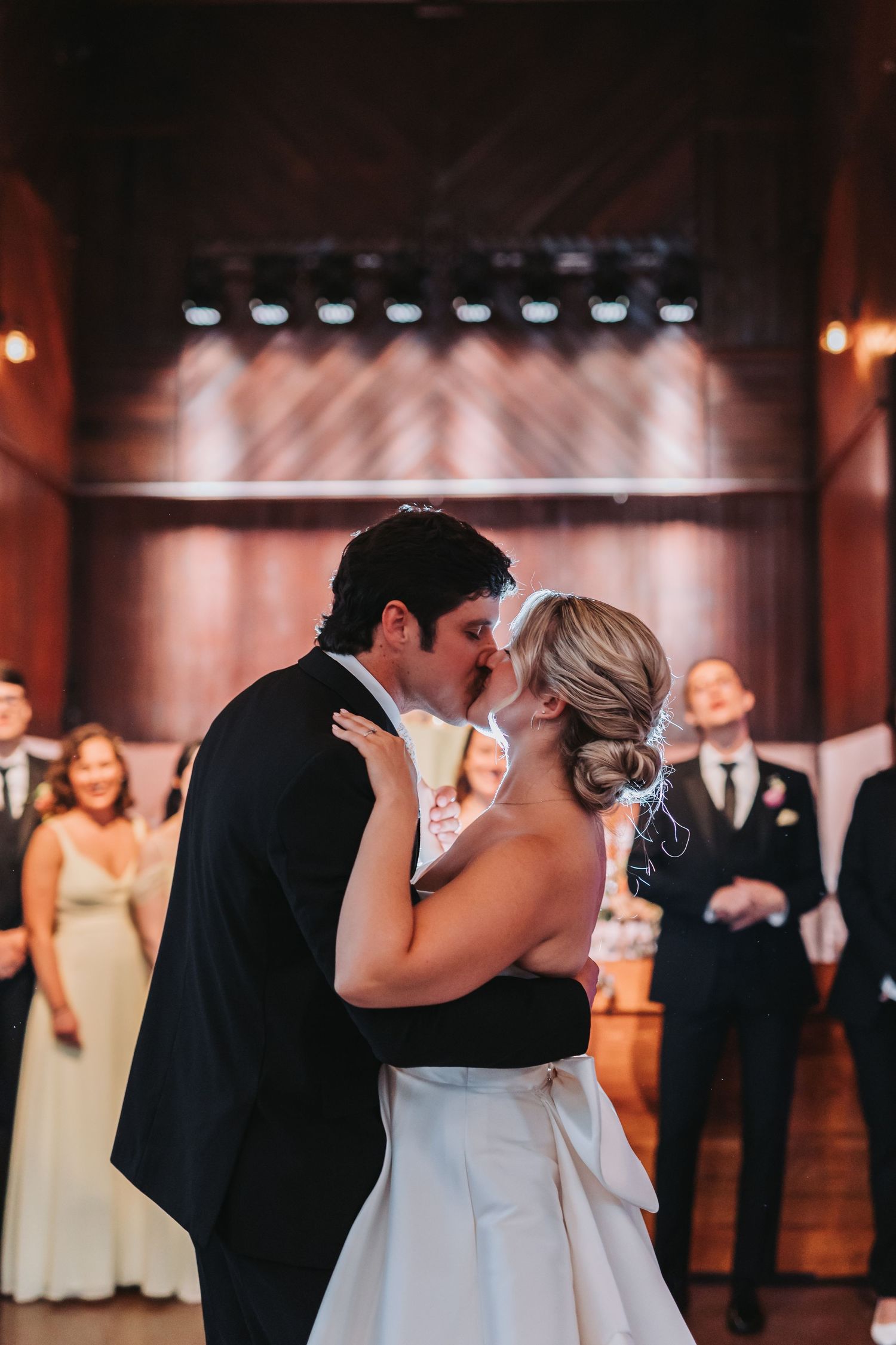Bride and groom share their first kiss on the dance floor surrounded by wedding guests at their reception.