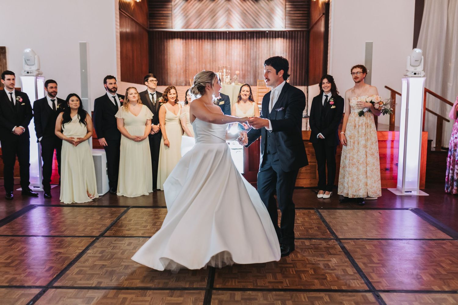 Bride and groom share first dance at wedding reception while bridal party looks on in elegant ballroom venue.
