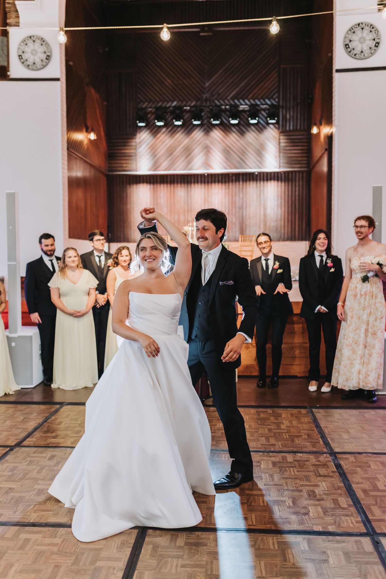Bride and groom share first dance as wedding guests watch in elegant wood-paneled reception hall.