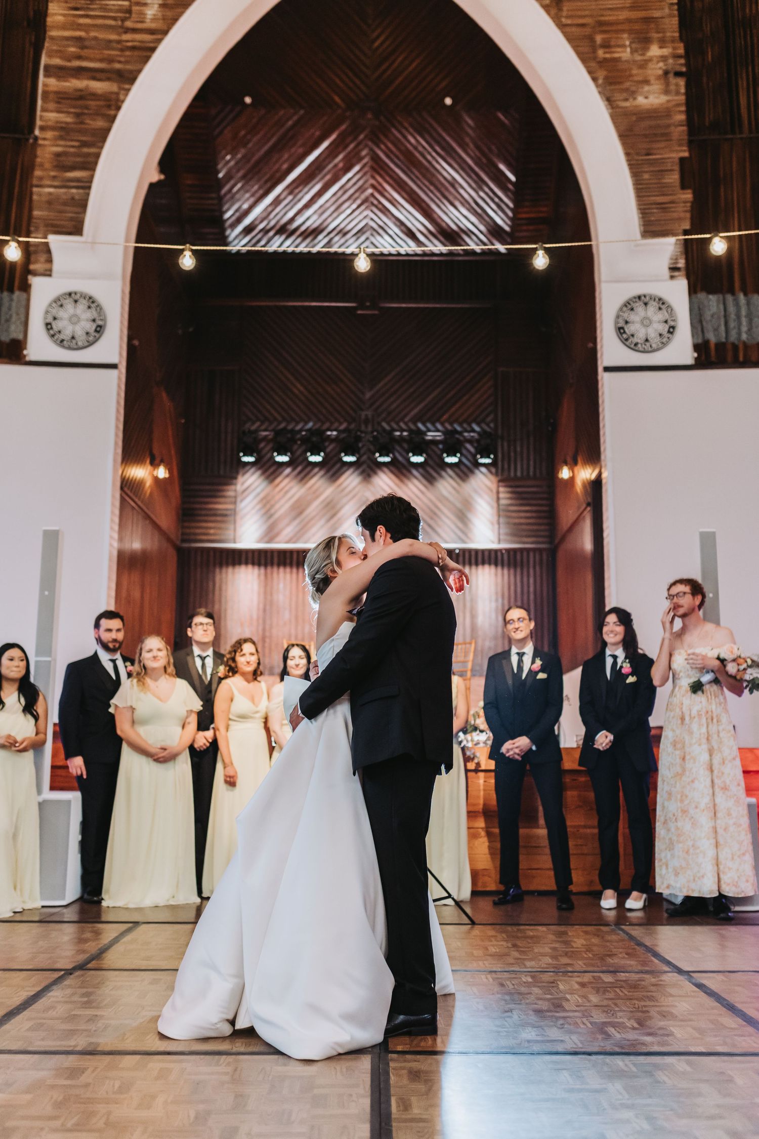 Bride and groom share first dance in stunning arched venue with wedding party watching.