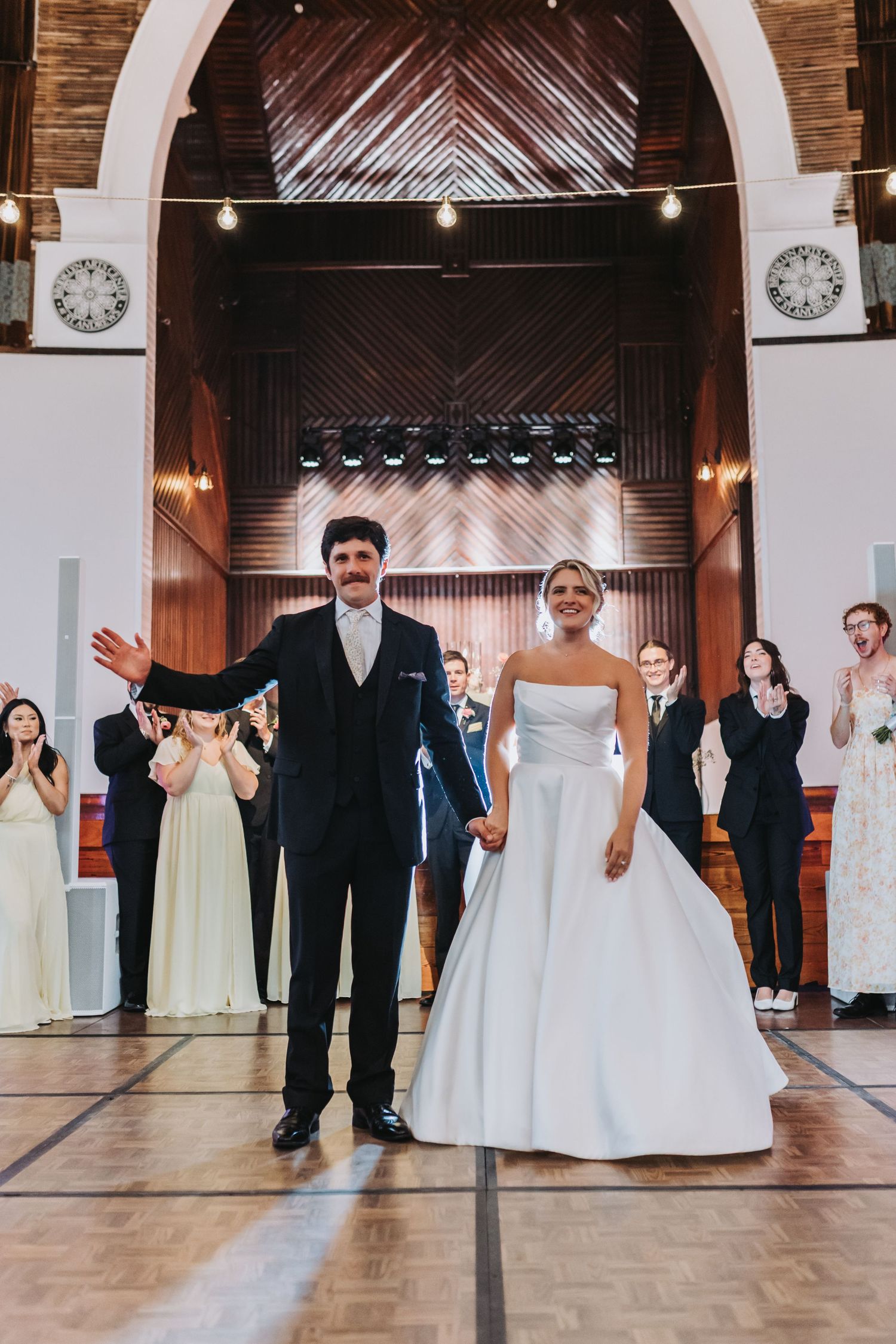 Bride and groom triumphantly enter their wedding reception to cheering guests in a grand hall.