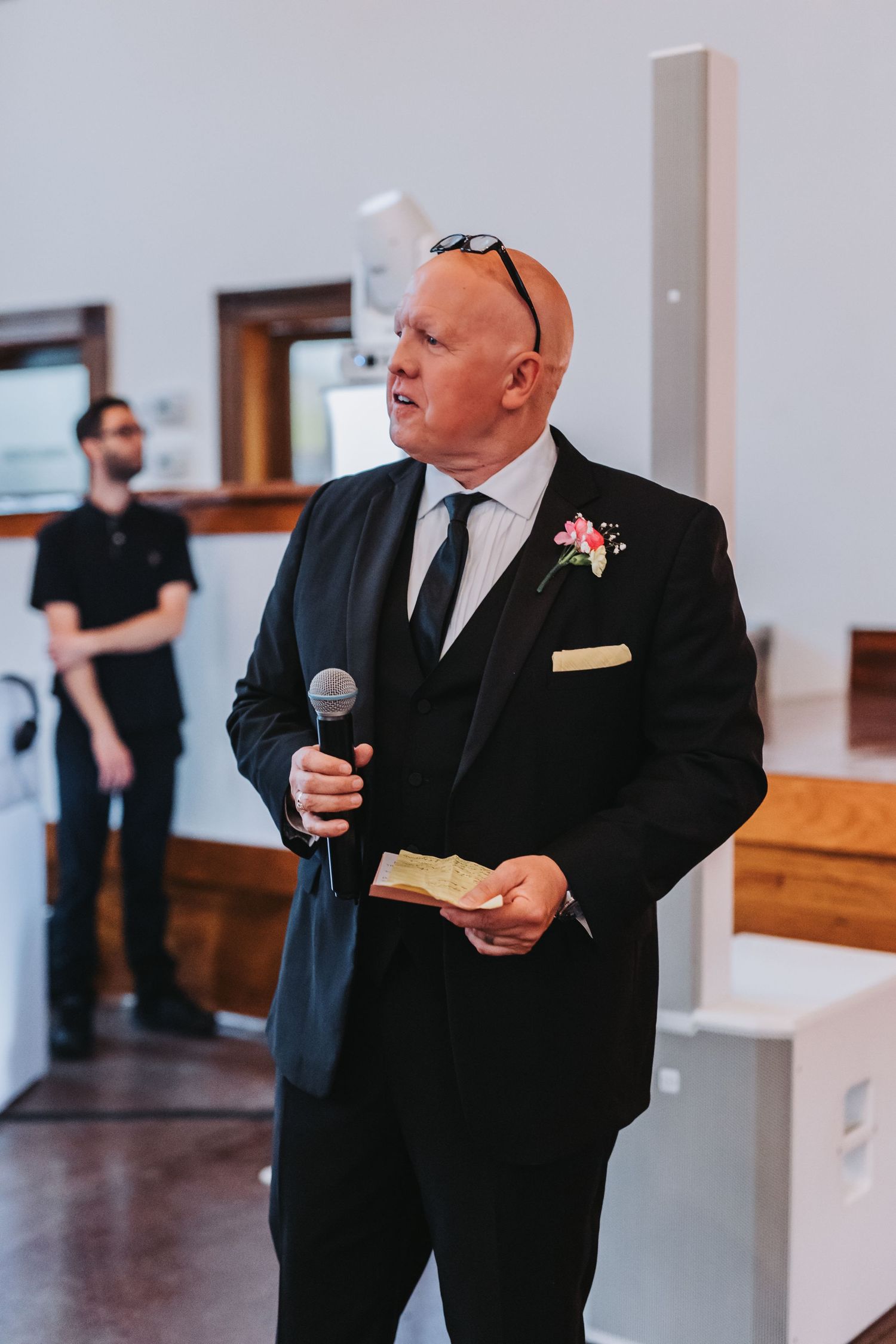 Man in black suit with boutonniere delivers a heartfelt wedding speech while holding a microphone.