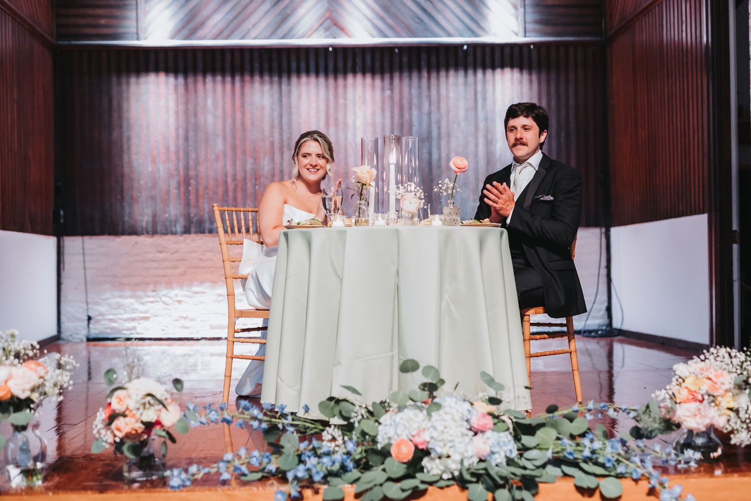 Bride and groom smiling at sweetheart table adorned with candles and floral arrangements at elegant wedding reception.