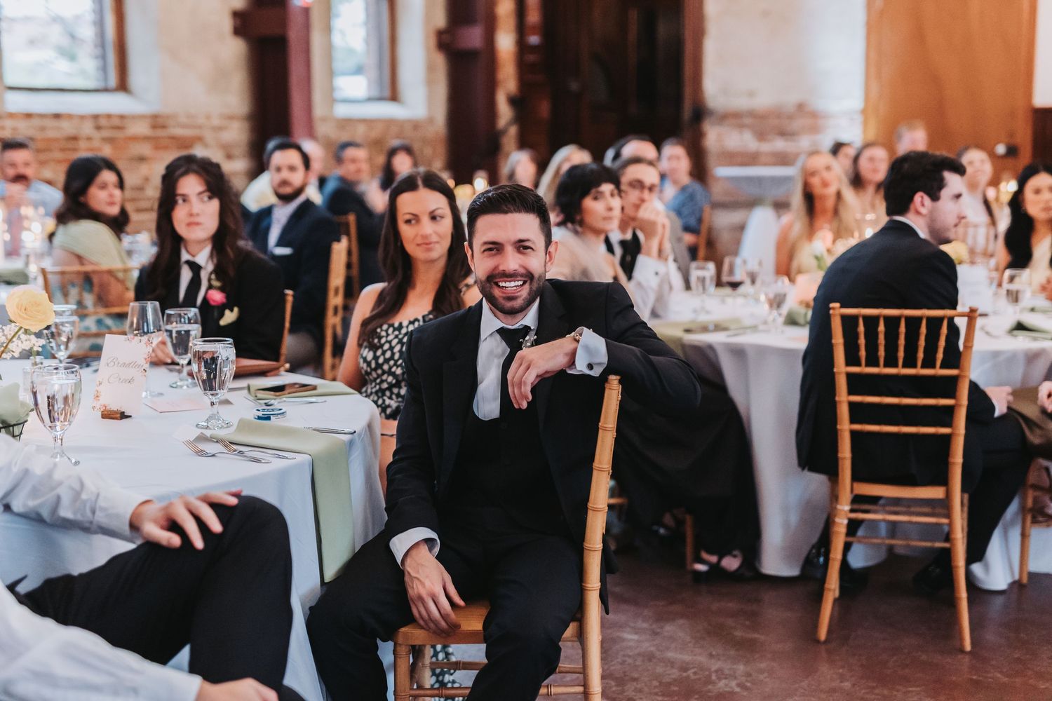 Smiling wedding guest in a black suit relaxes in a chiavari chair during wedding reception dinner.