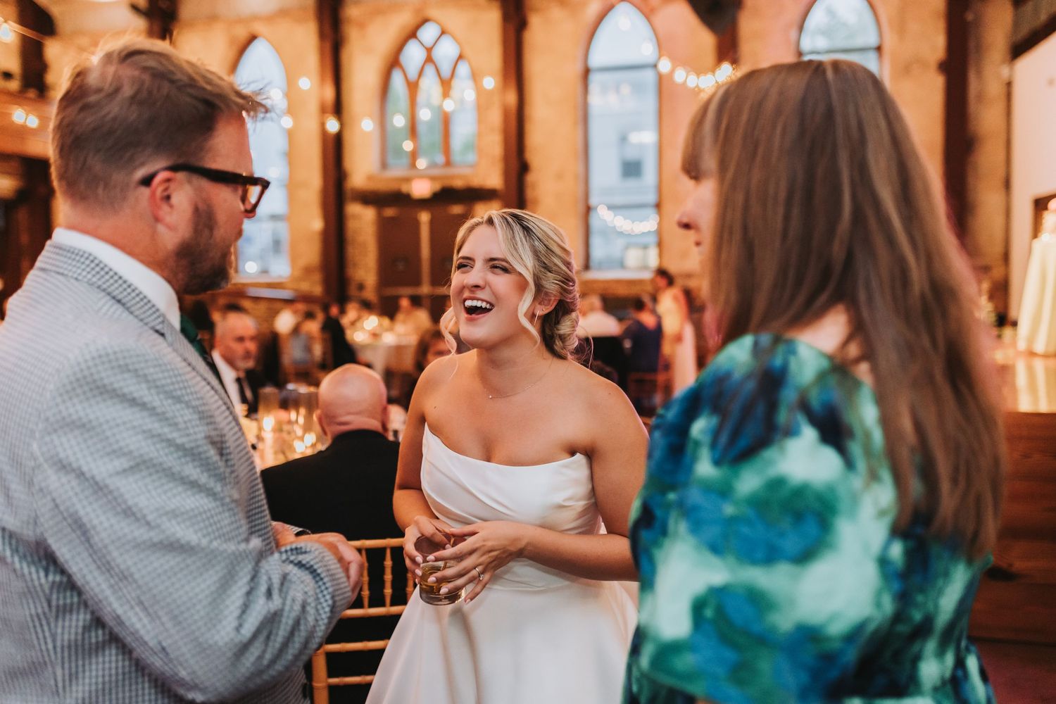 Smiling bride in strapless gown laughs with wedding guests inside a historic venue.