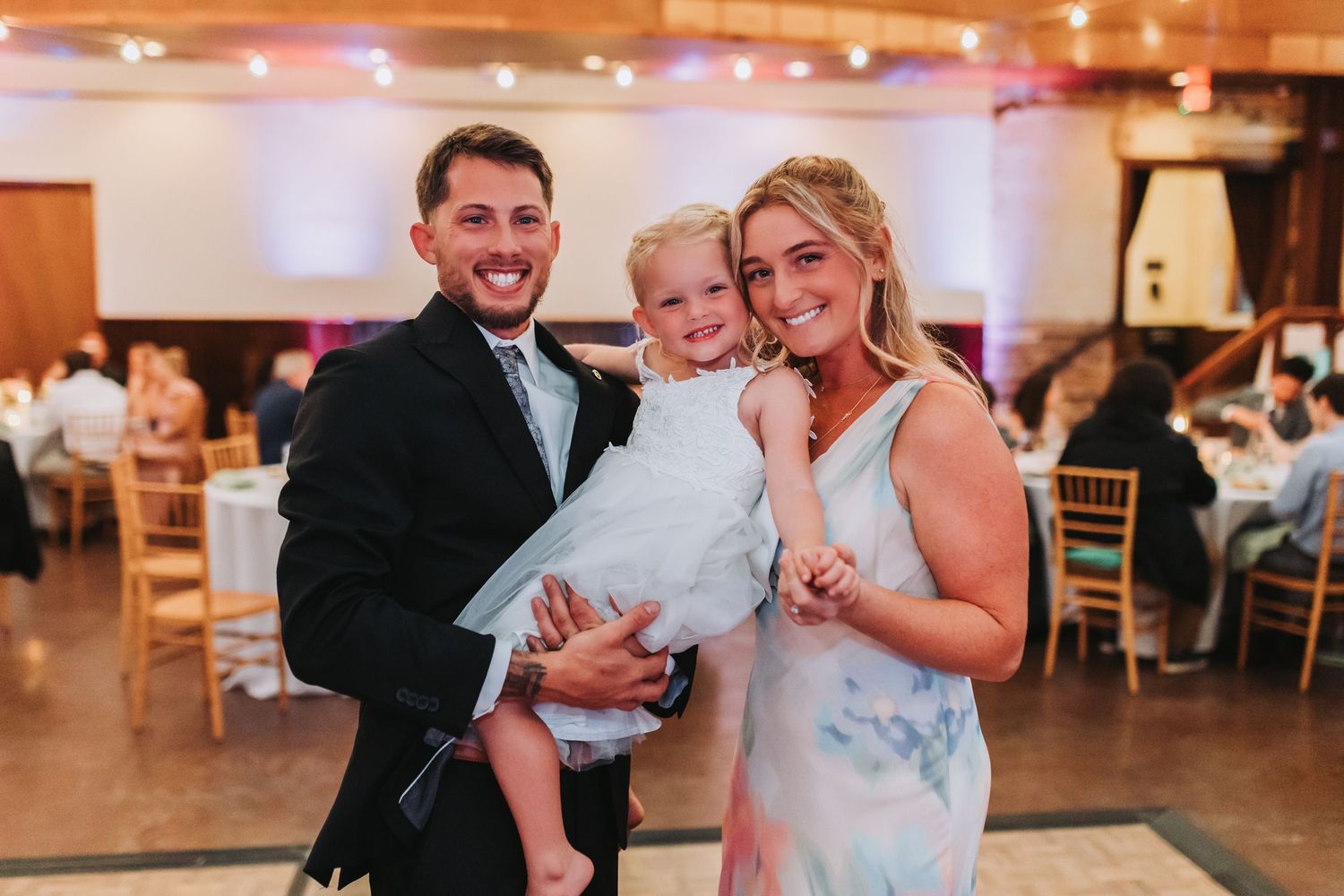 Smiling couple in formal wear holding a young child in white dress at an elegant wedding reception venue.