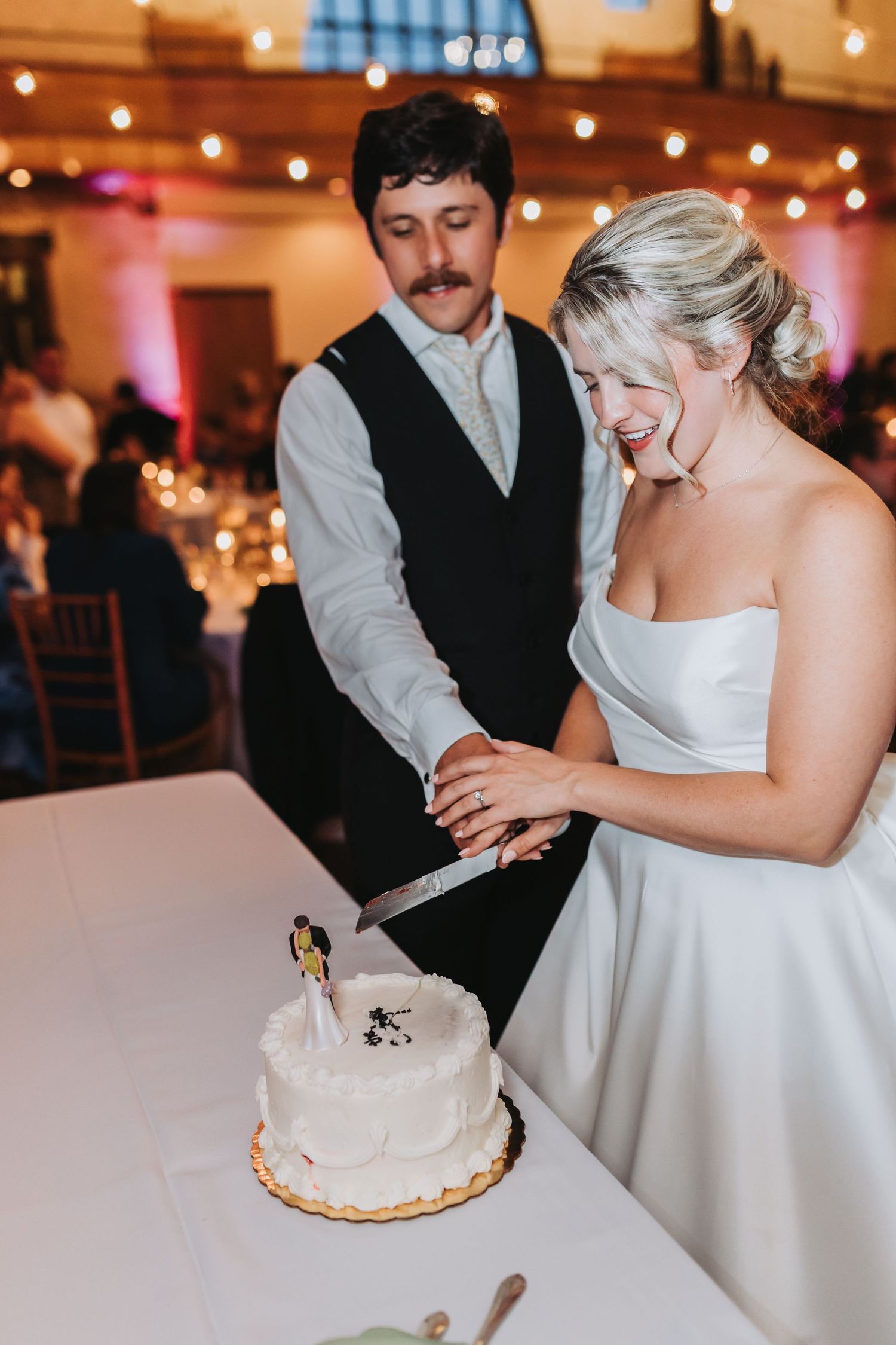 Bride and groom smiling while cutting their white two-tier wedding cake at their reception venue.