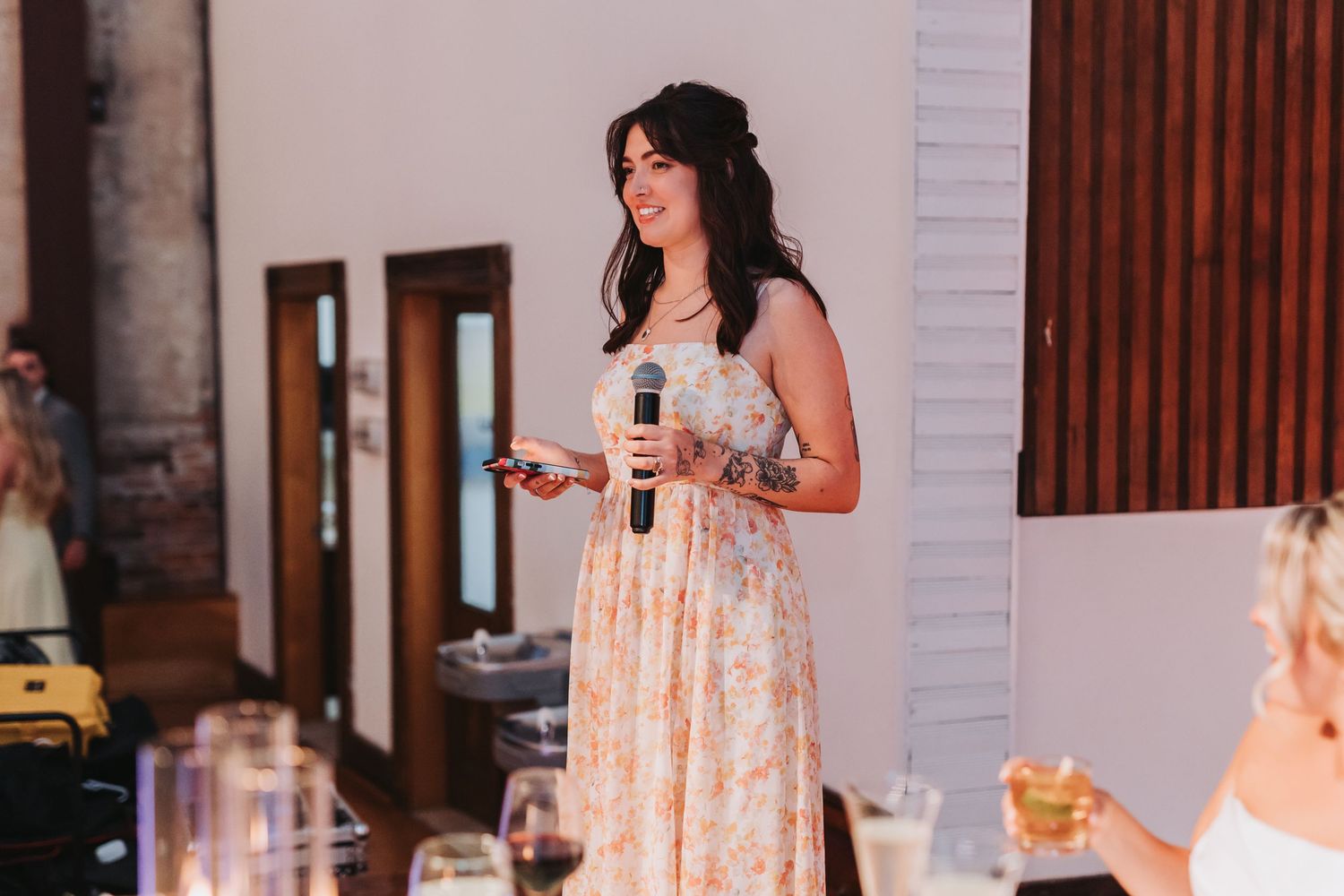 Woman in floral dress giving a speech with microphone at an elegant indoor wedding reception.