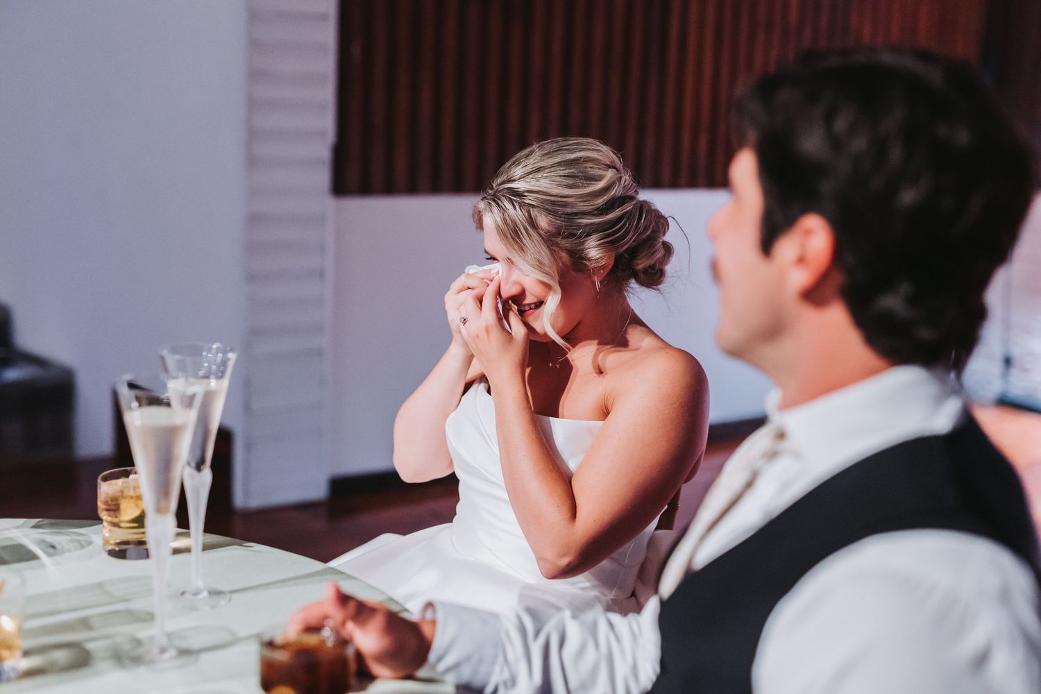 Emotional bride wiping tears at wedding reception table during heartfelt toasts.