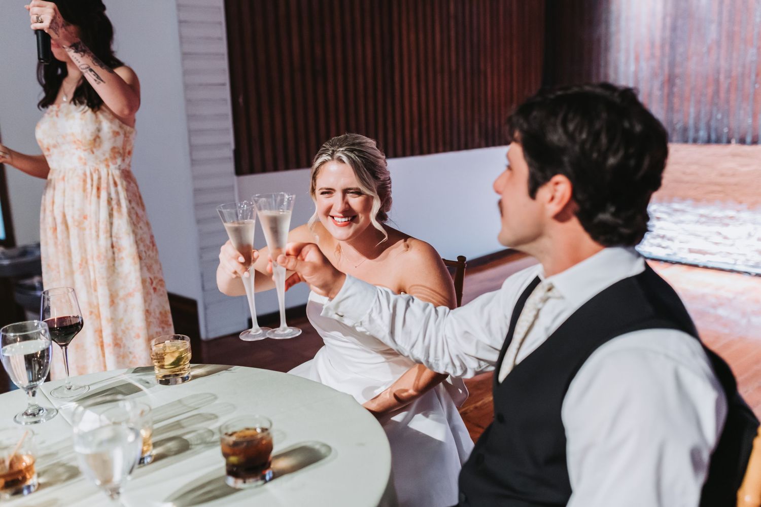 Bride and groom clinking champagne glasses at wedding reception table, smiling joyfully, with guests in background.