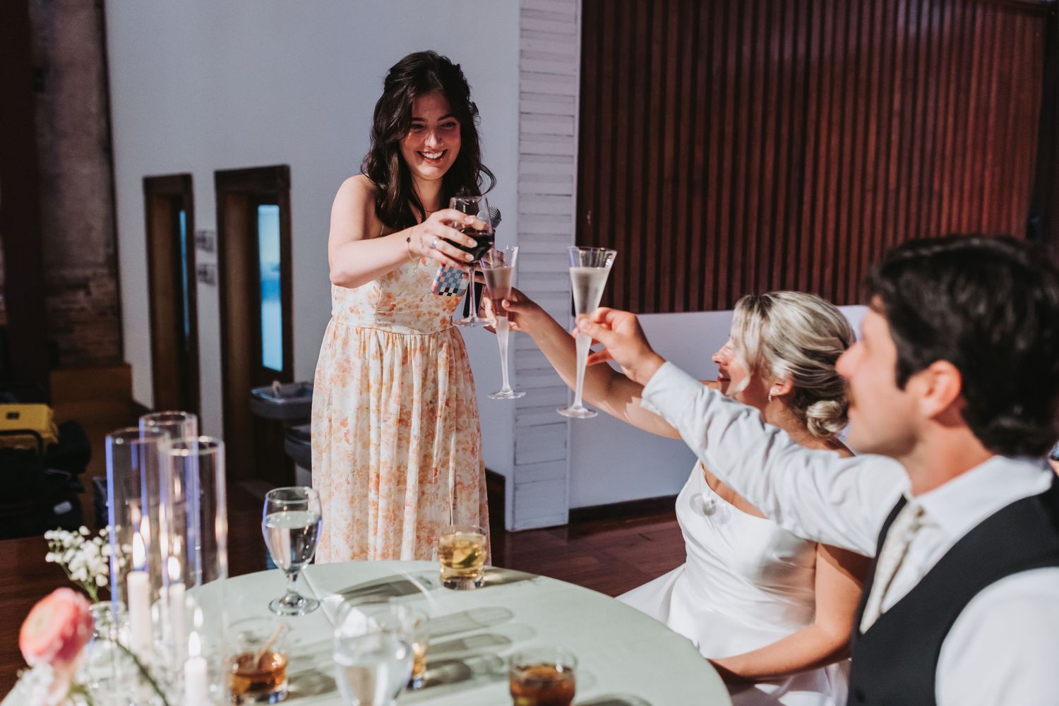 A woman in a floral dress toasts with wine glasses at an elegant wedding reception dinner table.