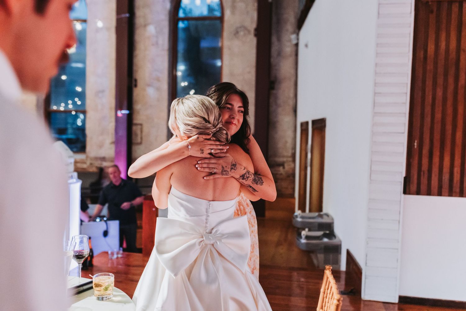 Bride shares a warm embrace with a tattooed guest during wedding reception celebration.