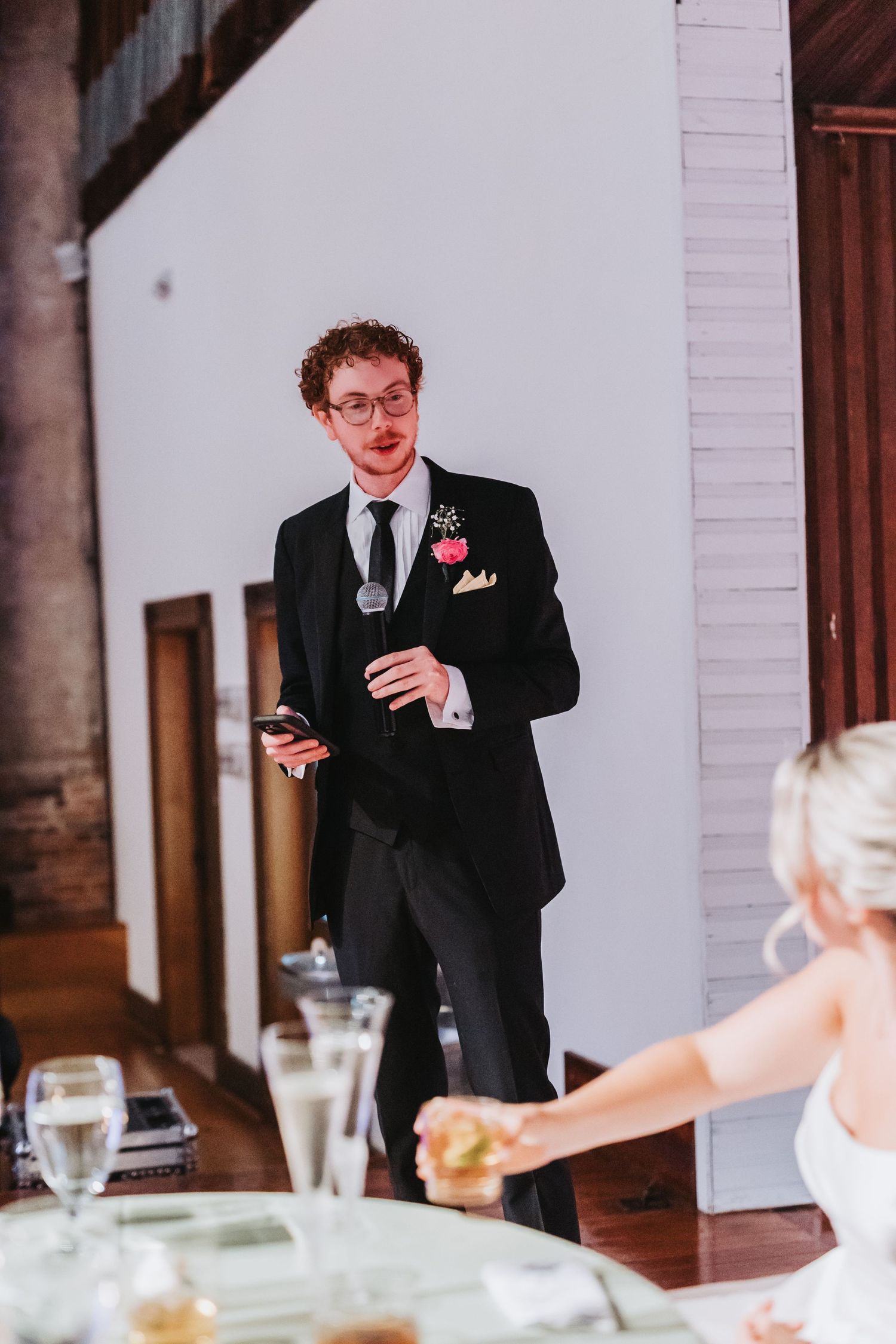 Groomsman in black tuxedo delivers wedding speech holding microphone and phone.