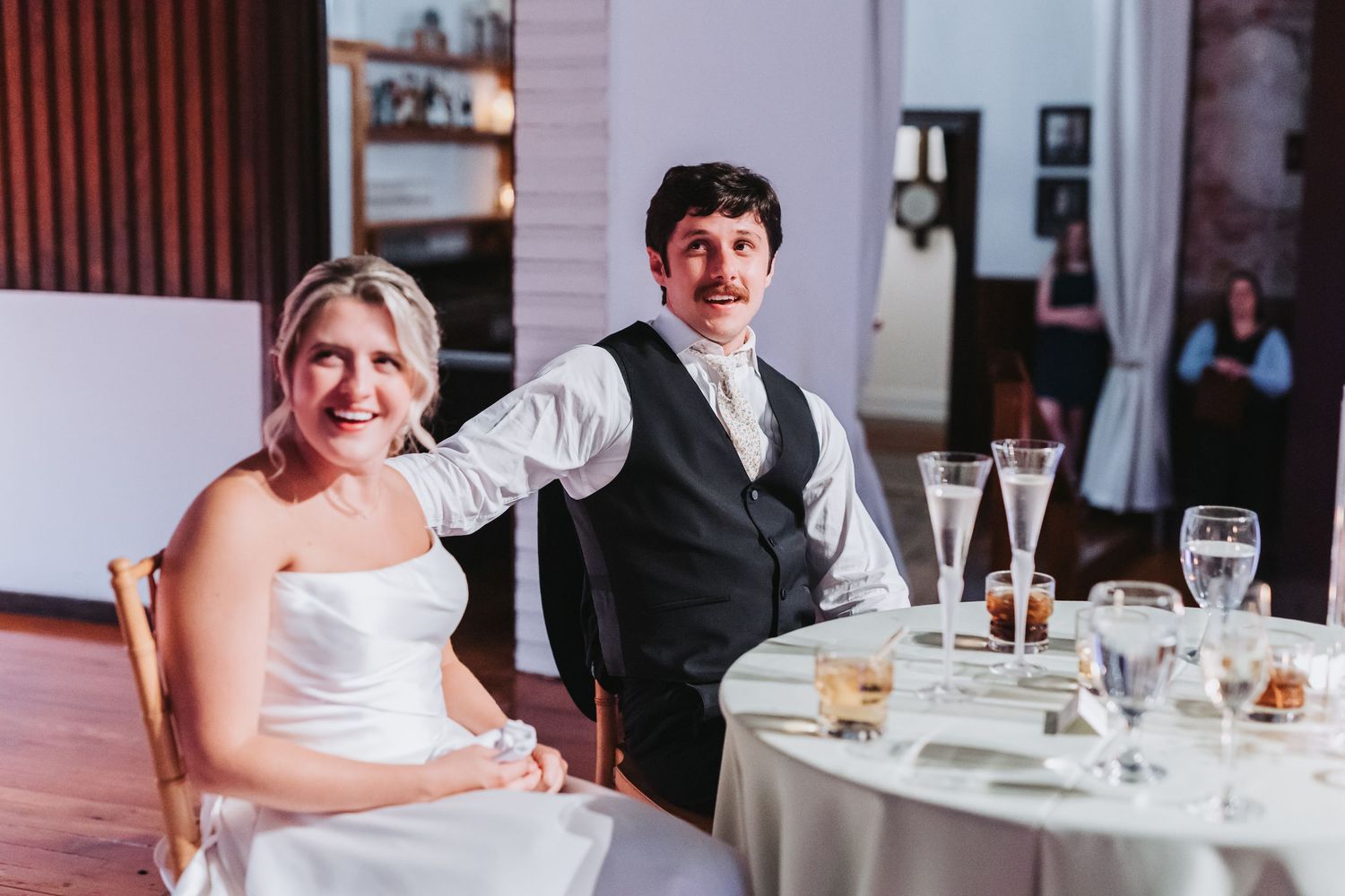 Bride and groom smile at sweetheart table during wedding reception indoors.